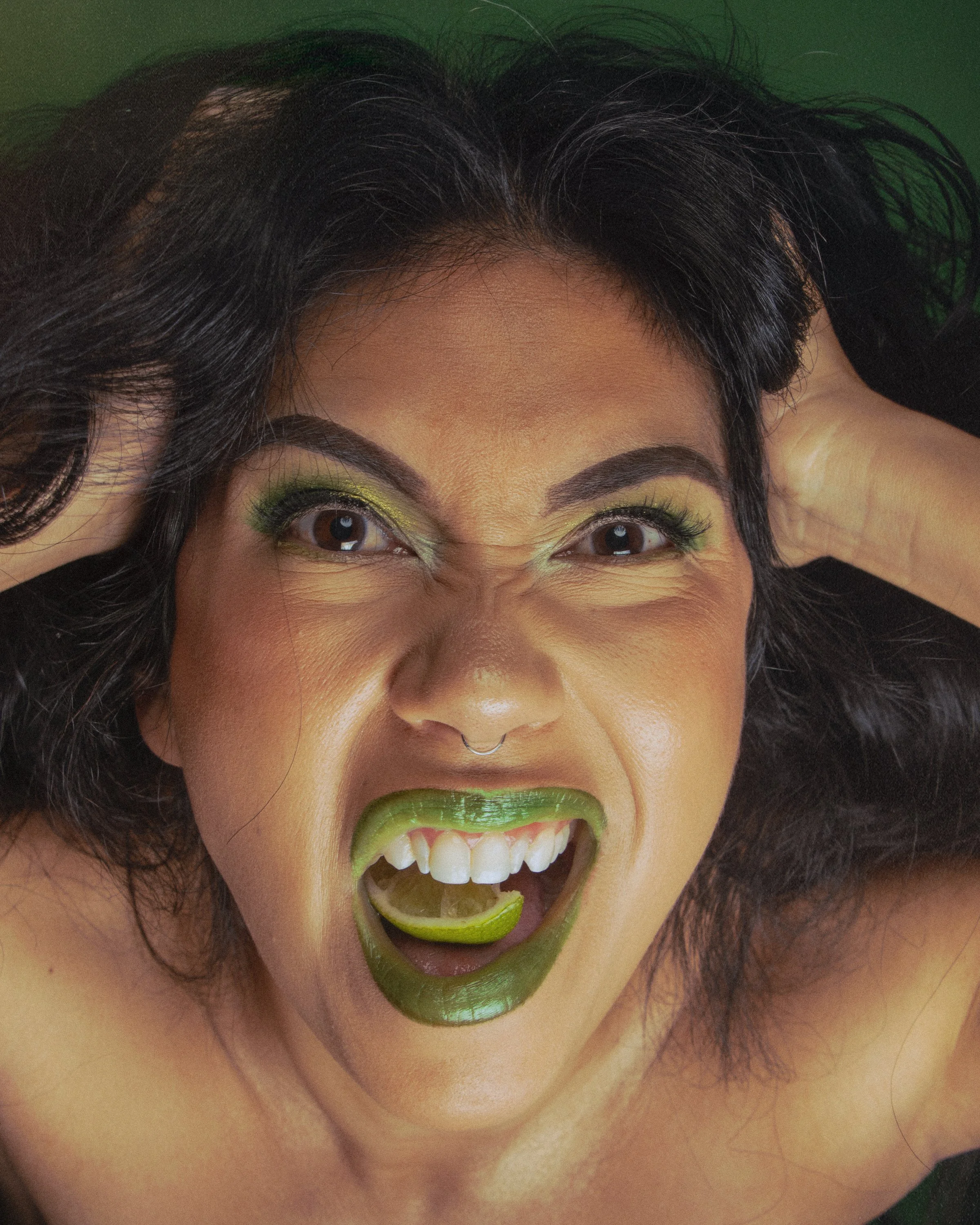 A woman with black curly hair and brown eyes, wearing vibrant green eyeshadow and lipstick, is smiling with her mouth open, showing her teeth and tongue, with her hands on her head.