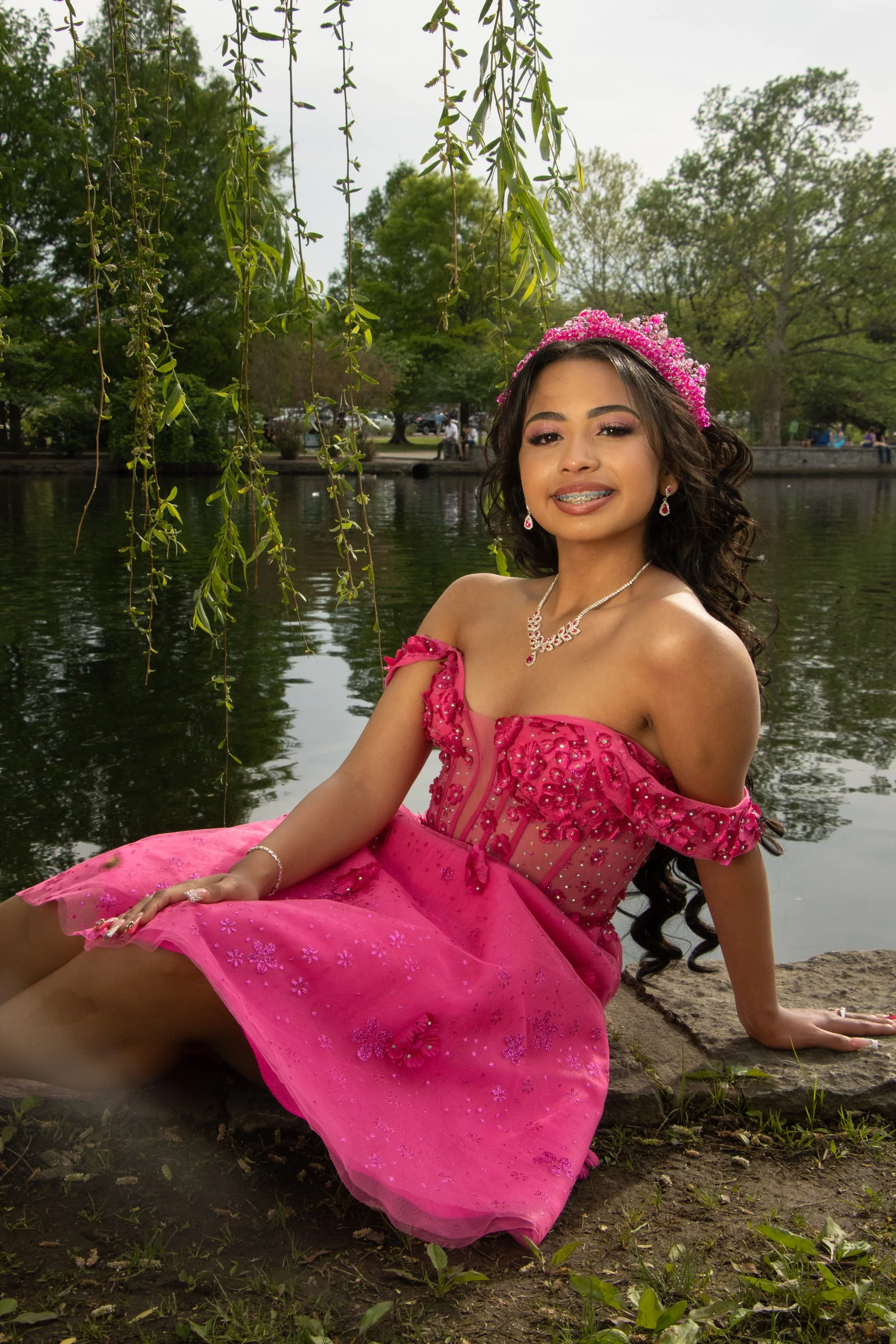A young woman in a pink dress sitting on the ground near a body of water, with hanging tree branches and lush greenery in the background.