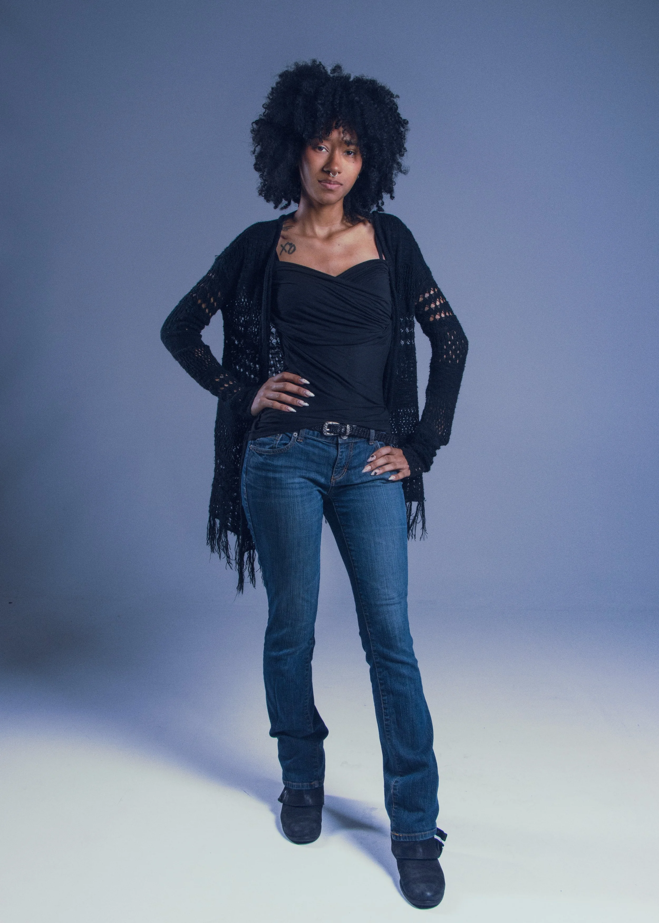 A woman with curly black hair, dressed in a black top, cardigan, and jeans, stands confidently with her hands on her hips against a plain background.
