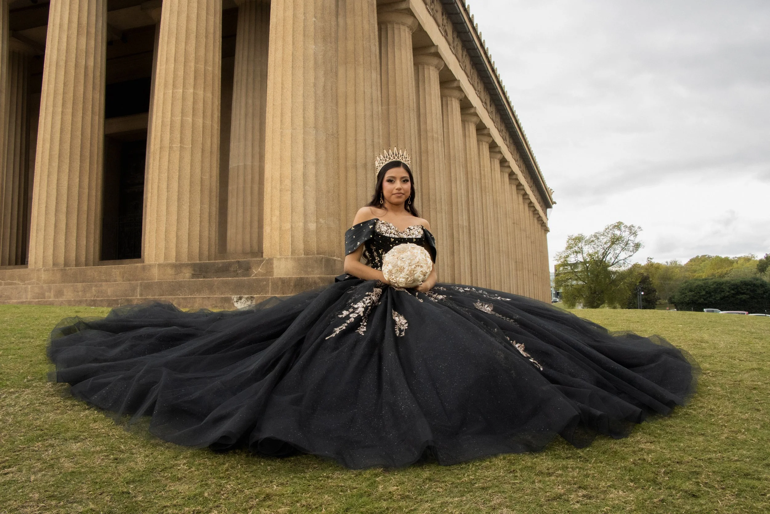 Young woman dressed in an elaborate black gown with floral embroidery, wearing a silver crown, sitting in front of the Lincoln Memorial, holding a bouquet of white roses, with overcast sky and trees in the background.