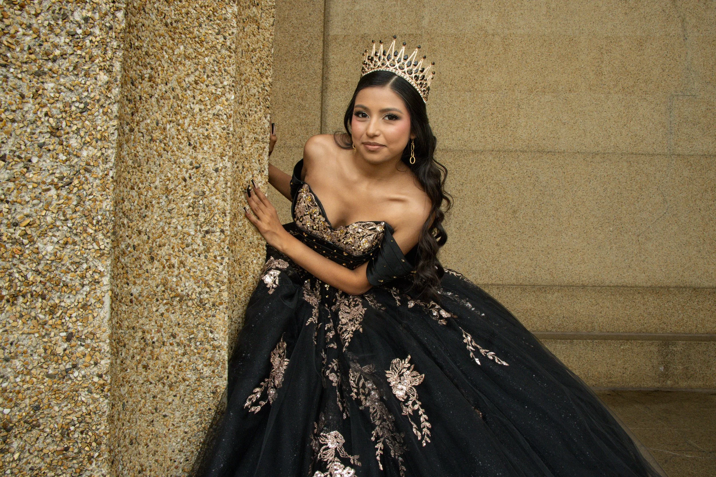 A woman wearing a crown and an elegant black and gold dress sits beside a textured wall, posing for the camera.