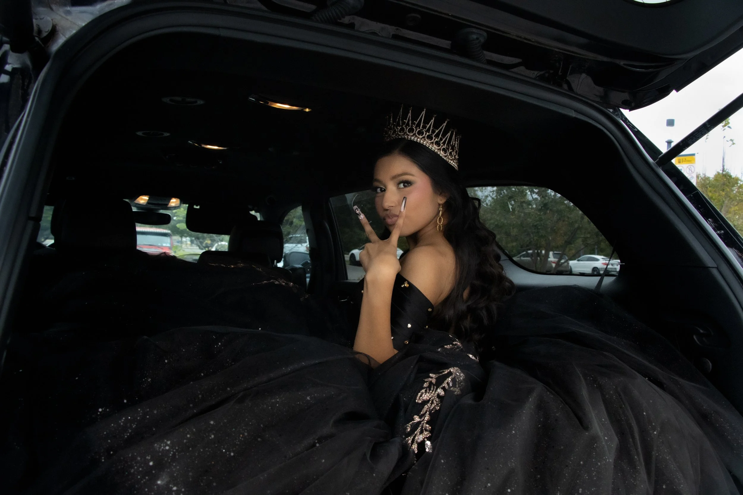 A woman wearing a tiara and black dress peeks out from inside a vehicle, with her tongue slightly out and holding up a peace sign gesture.