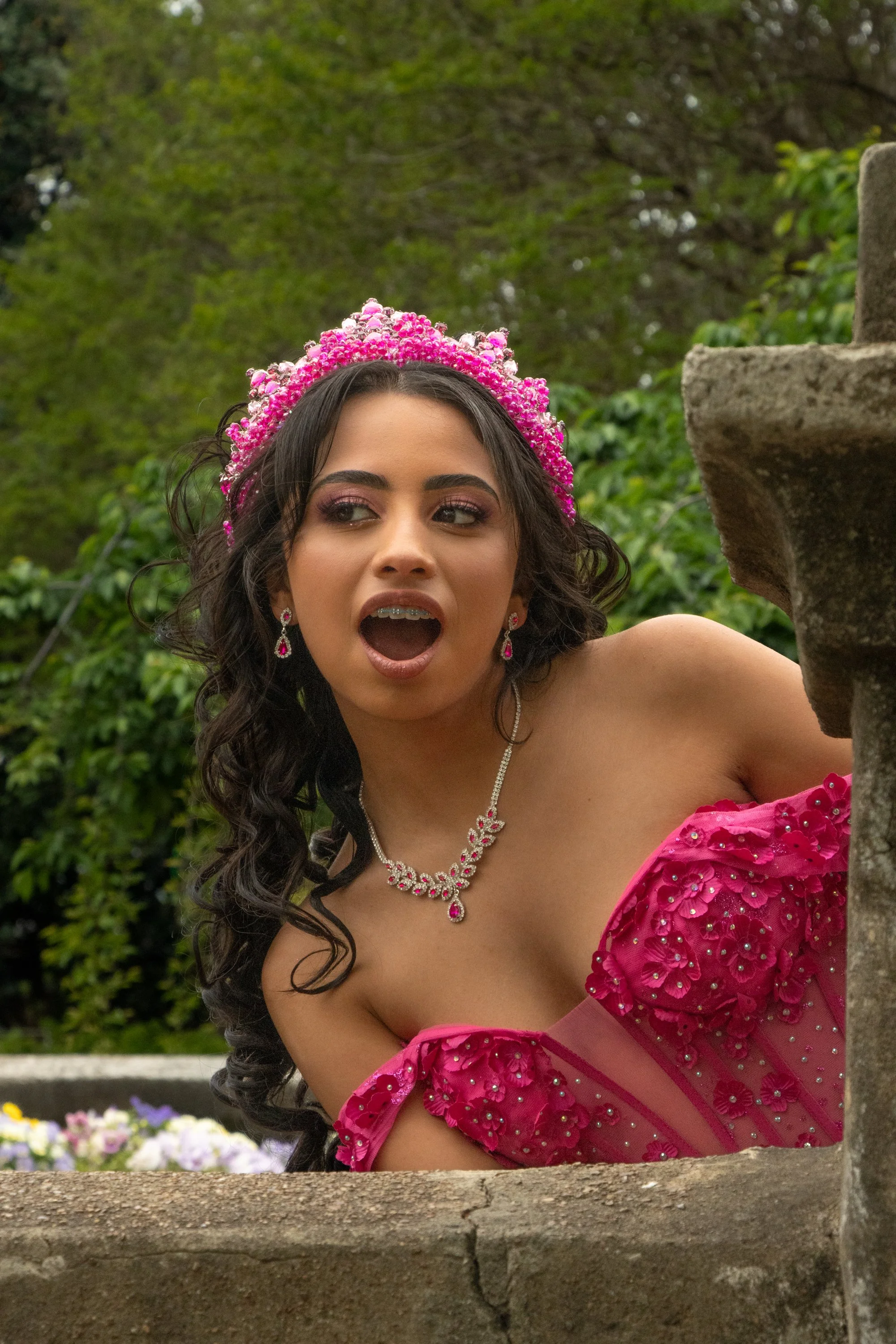 A woman in a pink dress with floral embellishments and jewelry, wearing a pink floral crown, appears to be leaning on a stone structure outdoors with greenery in the background.
