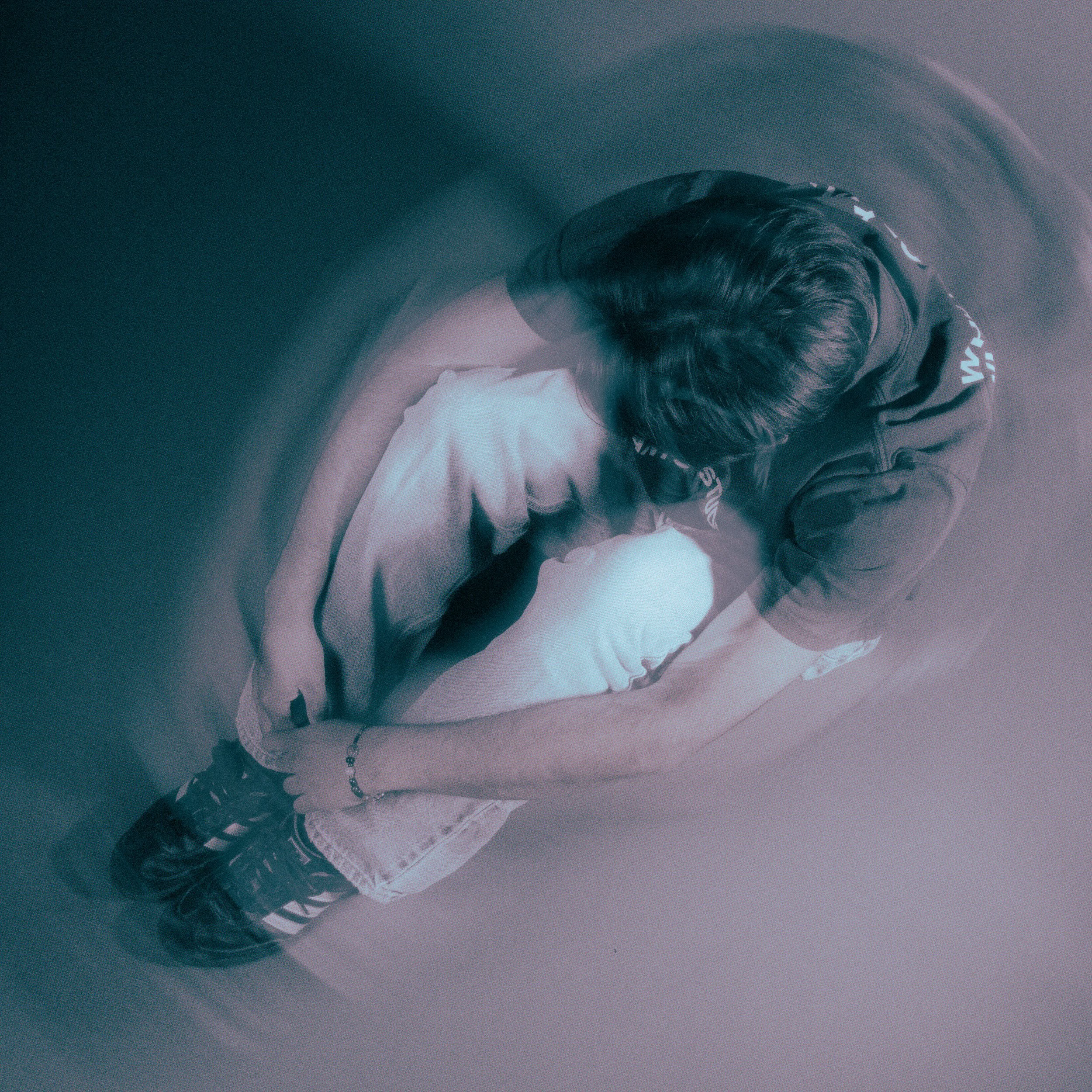 A person kneeling and leaning over a child who is lying on the floor, hugging their knees, in a dimly lit room.