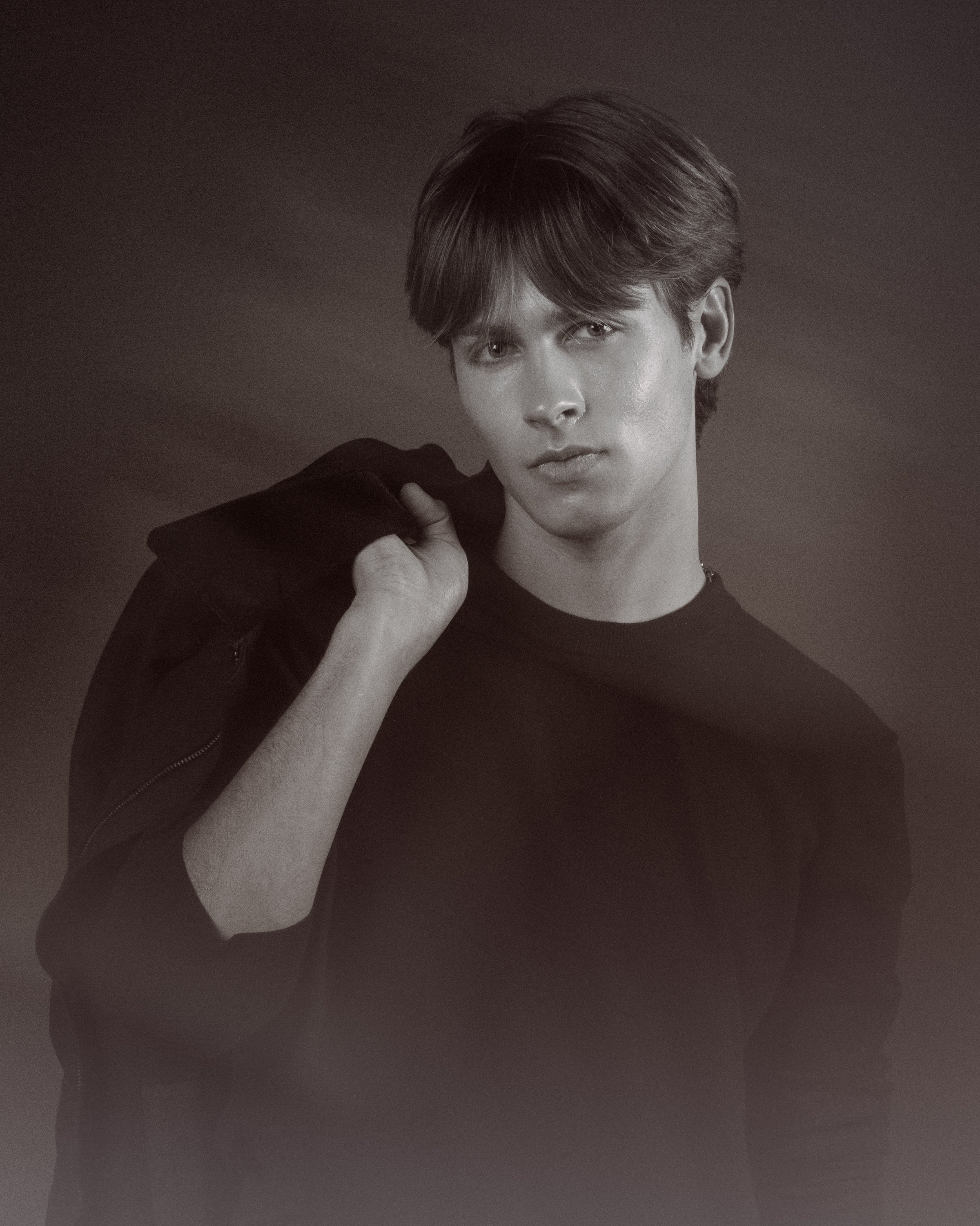 A young person with short, layered hair and a serious expression, wearing a dark top, holding a jacket over their shoulder, against a plain, dark background.
