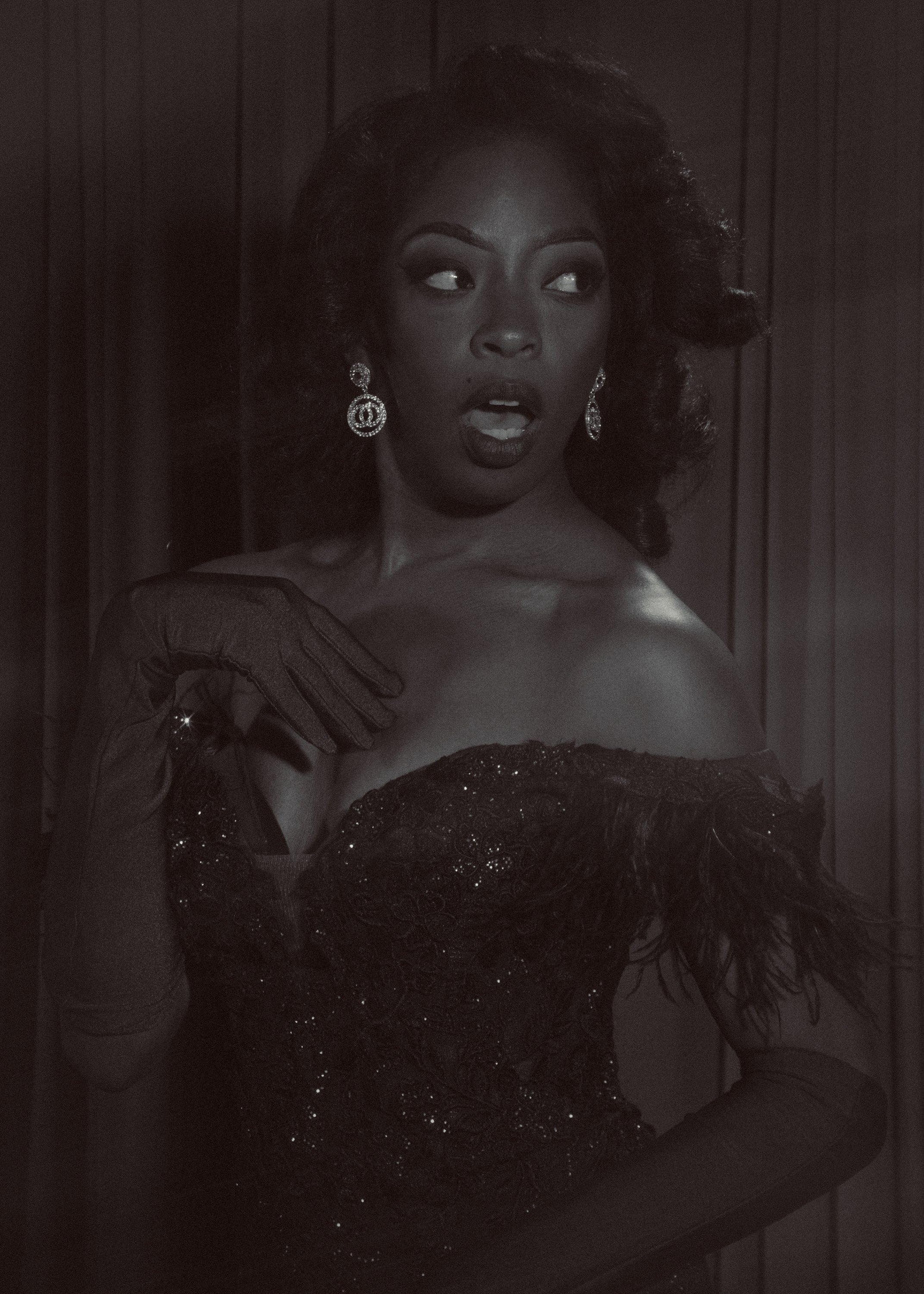 A woman with dark skin and voluminous hair styled in large curls is wearing elegant earrings and a strapless, black, embellished gown. She is posing in front of a dark, paneled background, with her mouth slightly open and eyes looking off to the side