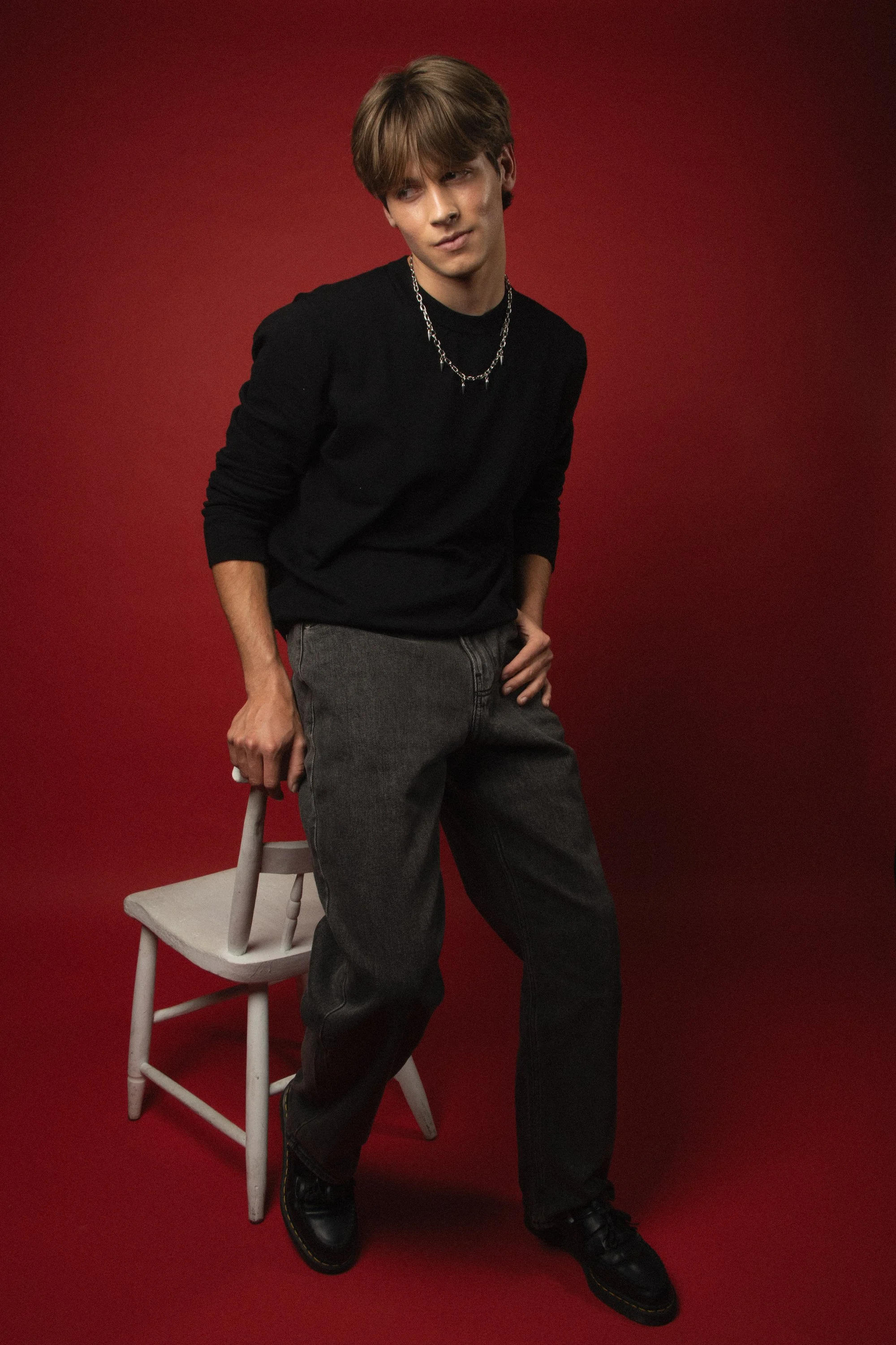 A young man with light brown hair wearing a black long-sleeve shirt and dark gray jeans, accessorized with a silver chain necklace, standing against a red background. He is resting one hand on a light-colored wooden chair, with a confident expression