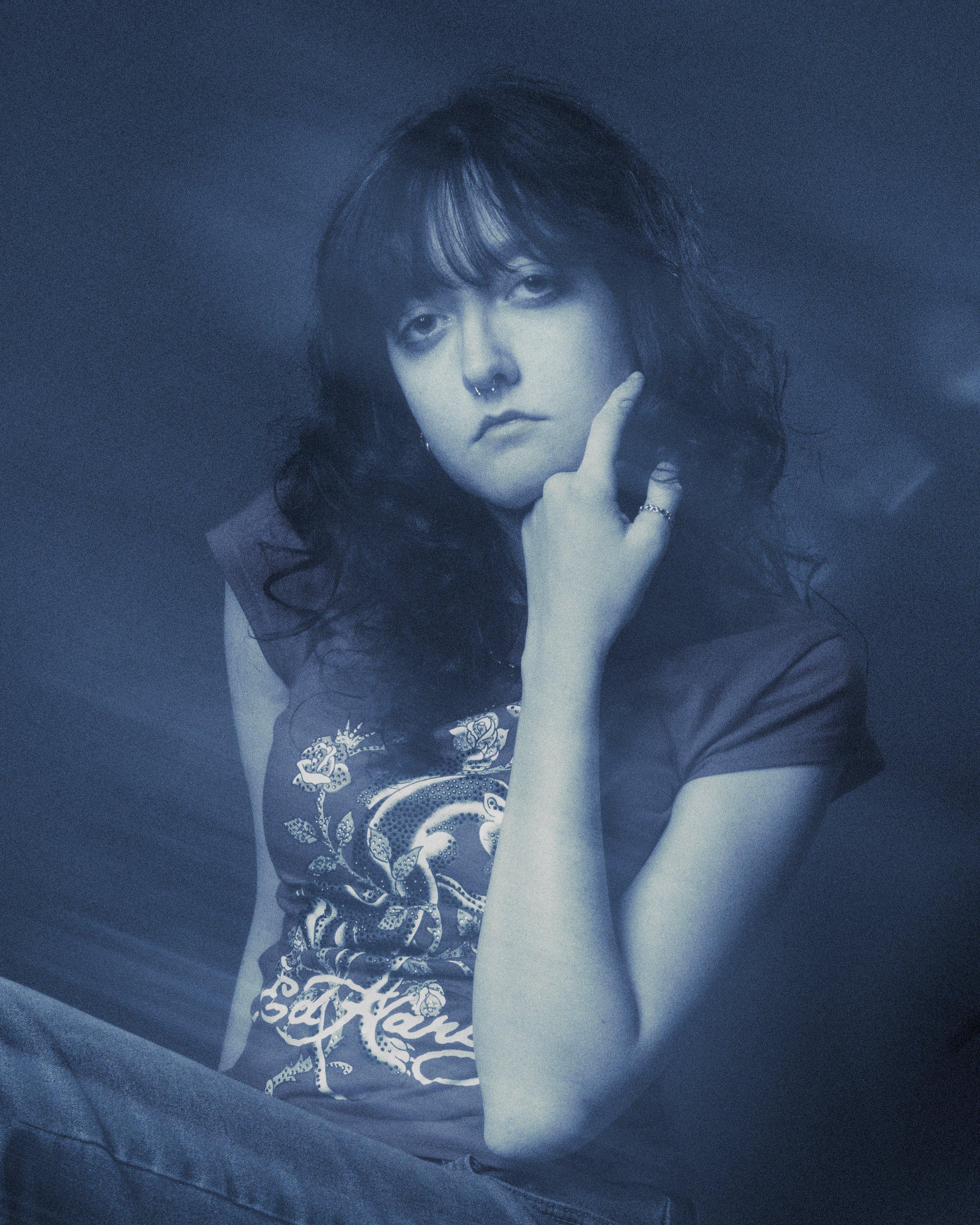 A person with long wavy hair and a septum piercing resting their chin on their hand, wearing a tattooed and graphic printed t-shirt, against a dark background.