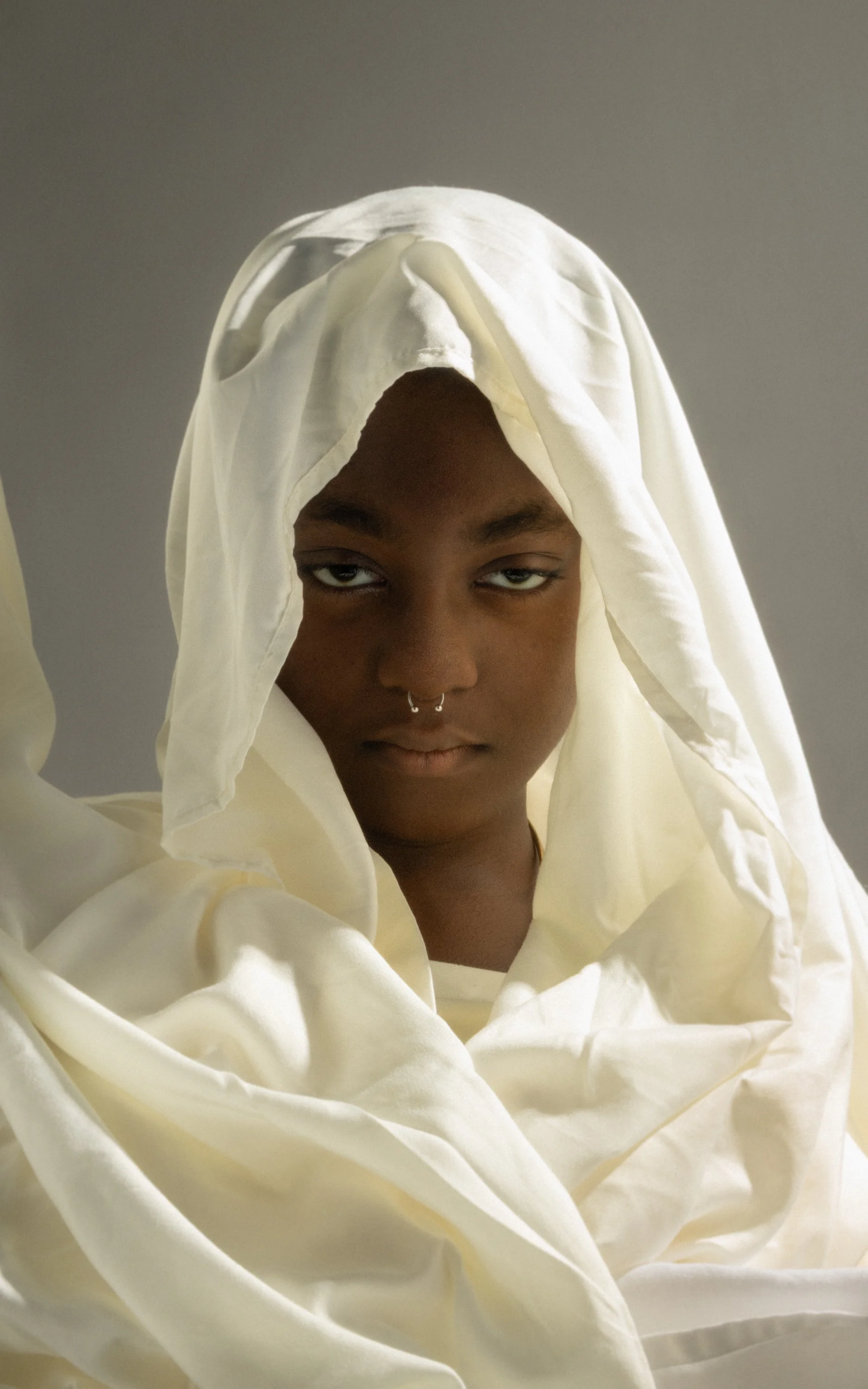A person with dark skin and dark eyes, wearing multiple jewelry piercings on their nose, is partially covered by draped, cream-colored fabric that surrounds their head and shoulders.