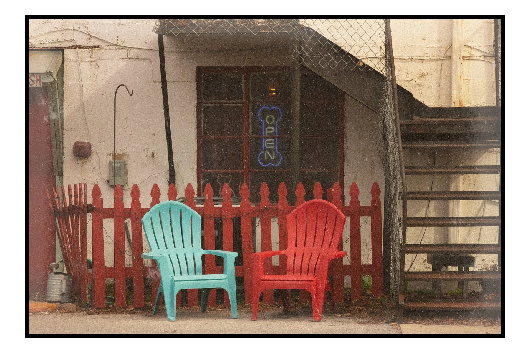 Two plastic Adirondack chairs, one teal and one red, are placed in front of a red picket fence. Behind the fence is a window with a neon sign that says 'Open' with a fishbone shape, and another light. To the right, a metal staircase leads upwards, an