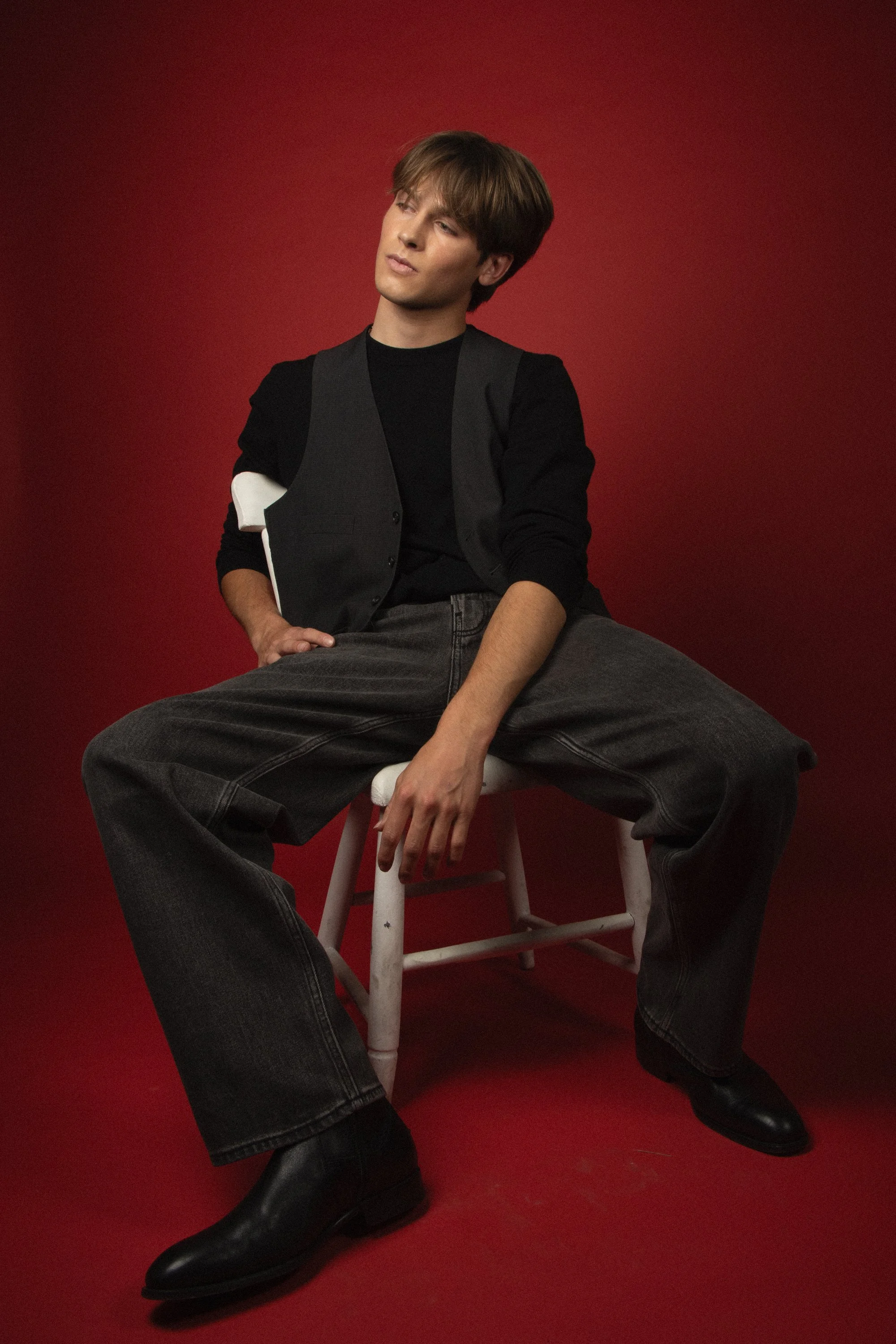 Young man with brown hair sitting on a stool in front of a red background, wearing a black shirt, gray vest, and large black boots, with legs spread wide apart and a serious expression.
