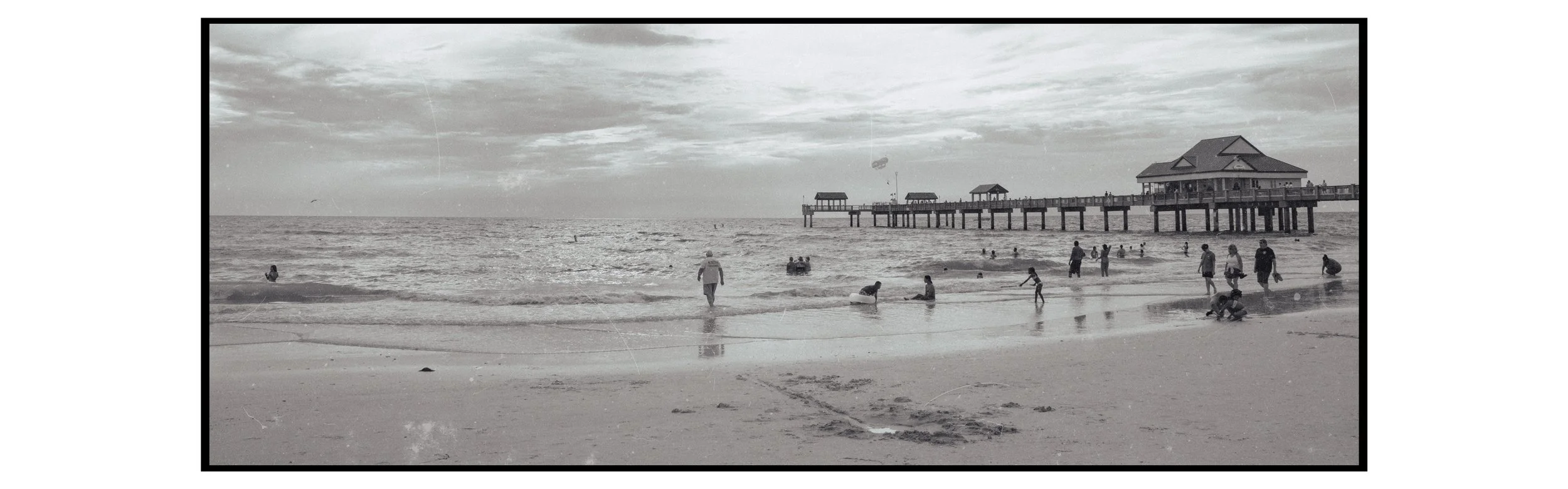 People enjoying a day at the beach with a pier extending into the water, some in the shallow waves and some further out, under a cloudy sky.