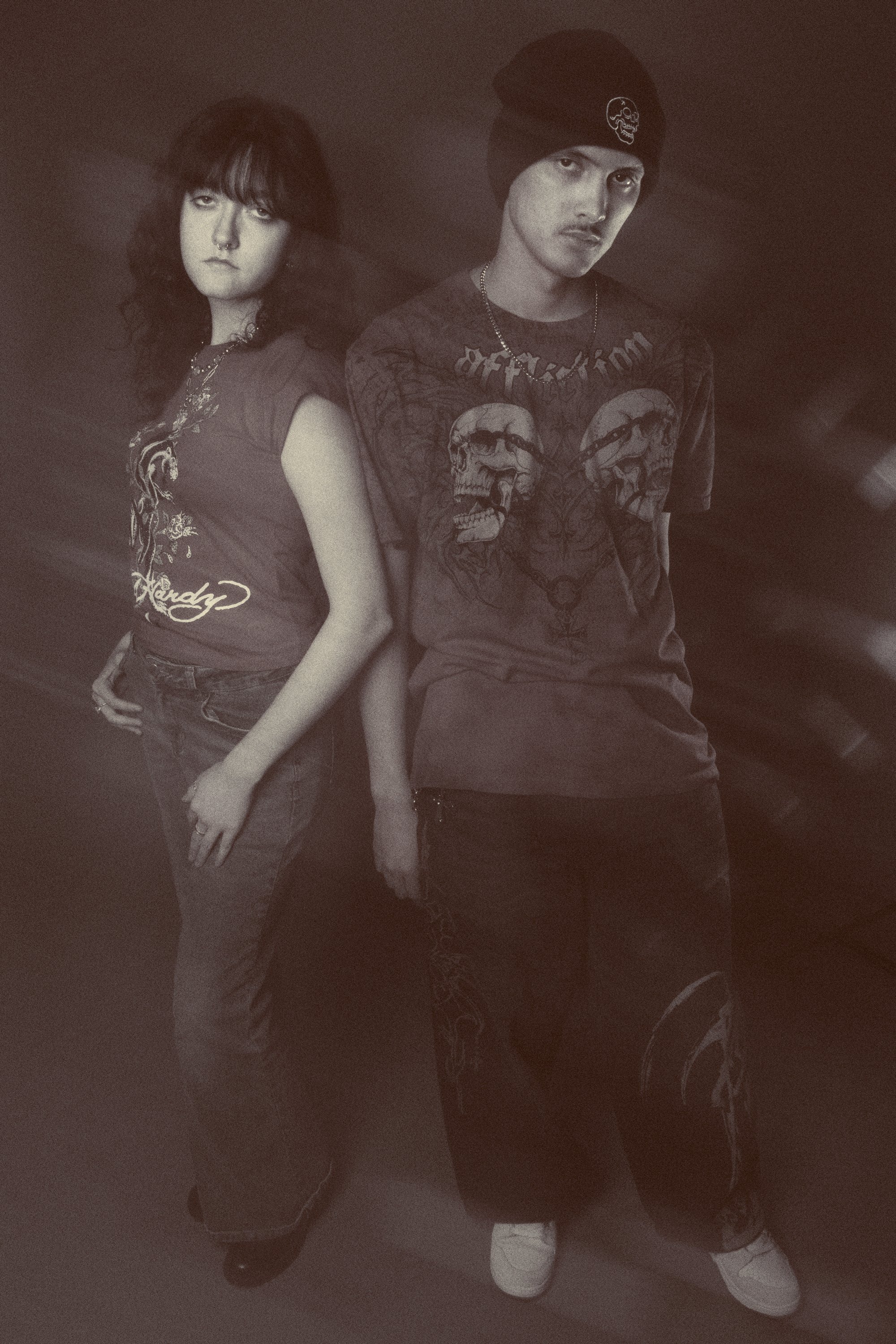 Two young adults with edgy fashion and serious expressions standing against a dark background. The person on the left has curly hair, a septum piercing, and is wearing a t-shirt with graphic design. The person on the right has straight hair, a beanie