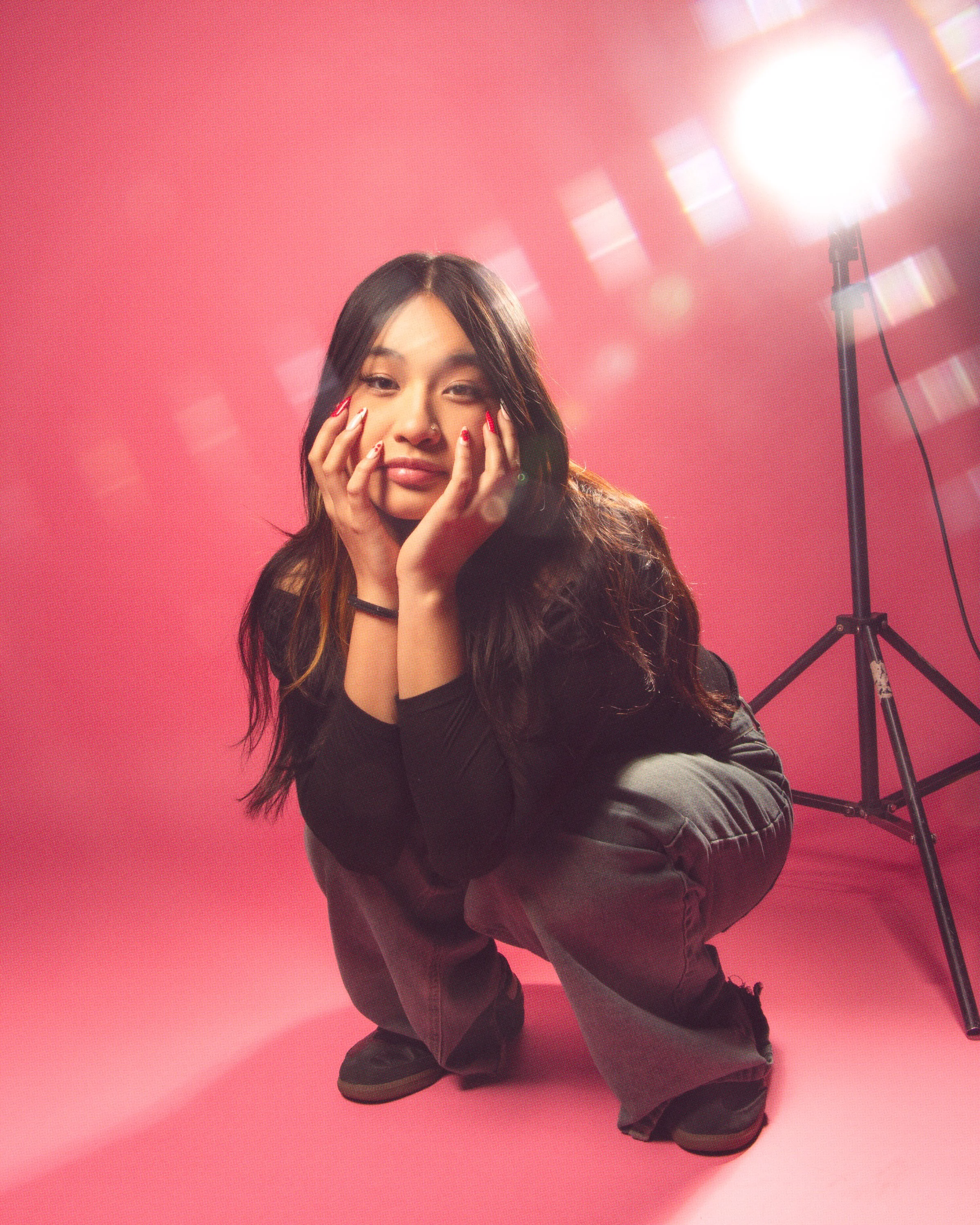 A woman with long dark hair posing with her hands on her face against a pink background, lit by a large studio light on a stand to her right.