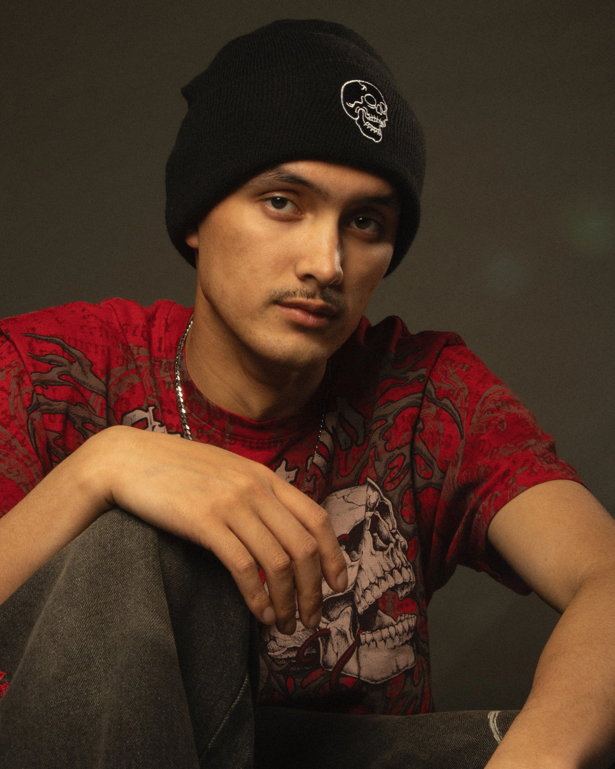 A young man wearing a black beanie with a skull design, a red graphic T-shirt featuring a skull, and sitting indoors with a gray background. His right arm is resting on his leg and he is looking at the camera.