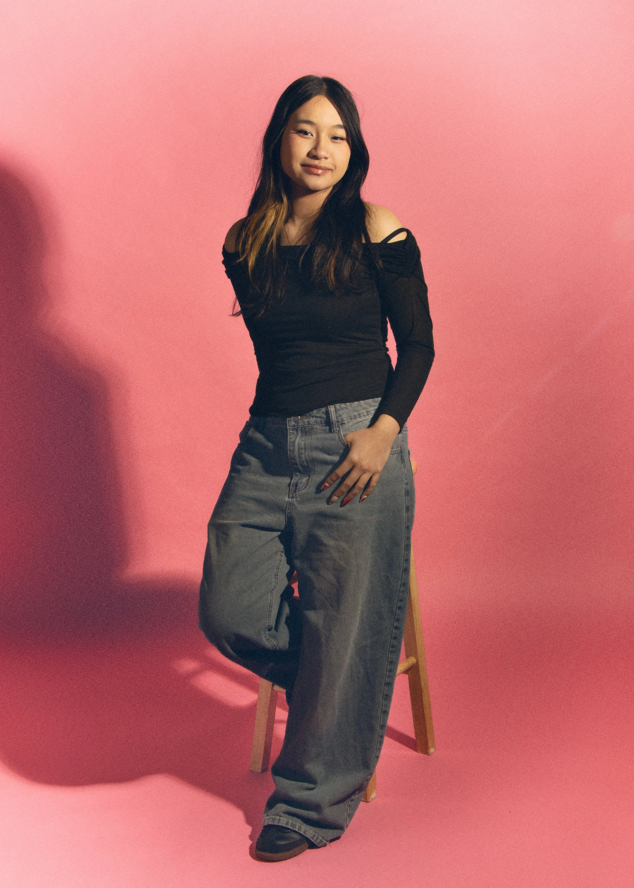Young woman with long dark hair standing against a pink background, wearing a black off-the-shoulder top and loose gray jeans, with one hand in her pocket and smiling.