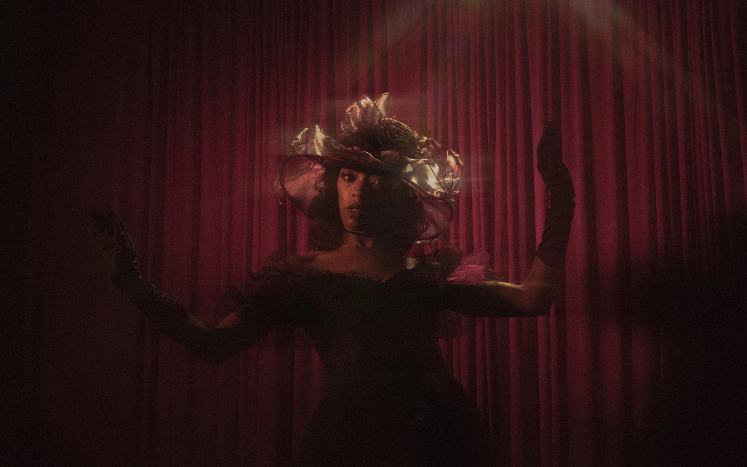 A woman wearing a large, colorful hat with flower and feather details, dressed in dark clothing, poses with her arms raised against a backdrop of red curtains.
