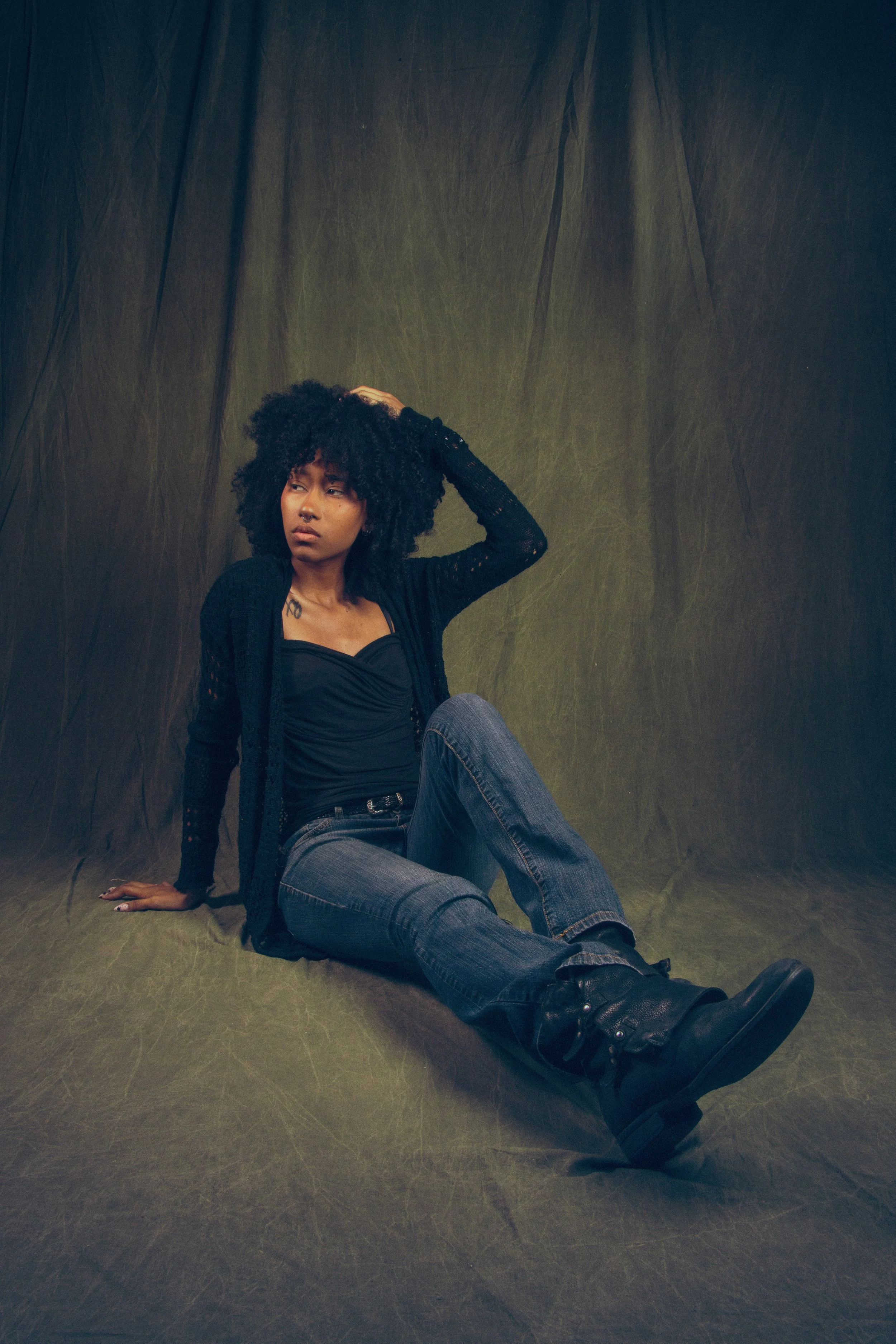 A woman with curly black hair sitting on the floor against a dark green backdrop. She is wearing a black top, jeans, black boots, and a black cardigan. She has a tattoo on her chest and is looking to the side with one hand resting on her head.