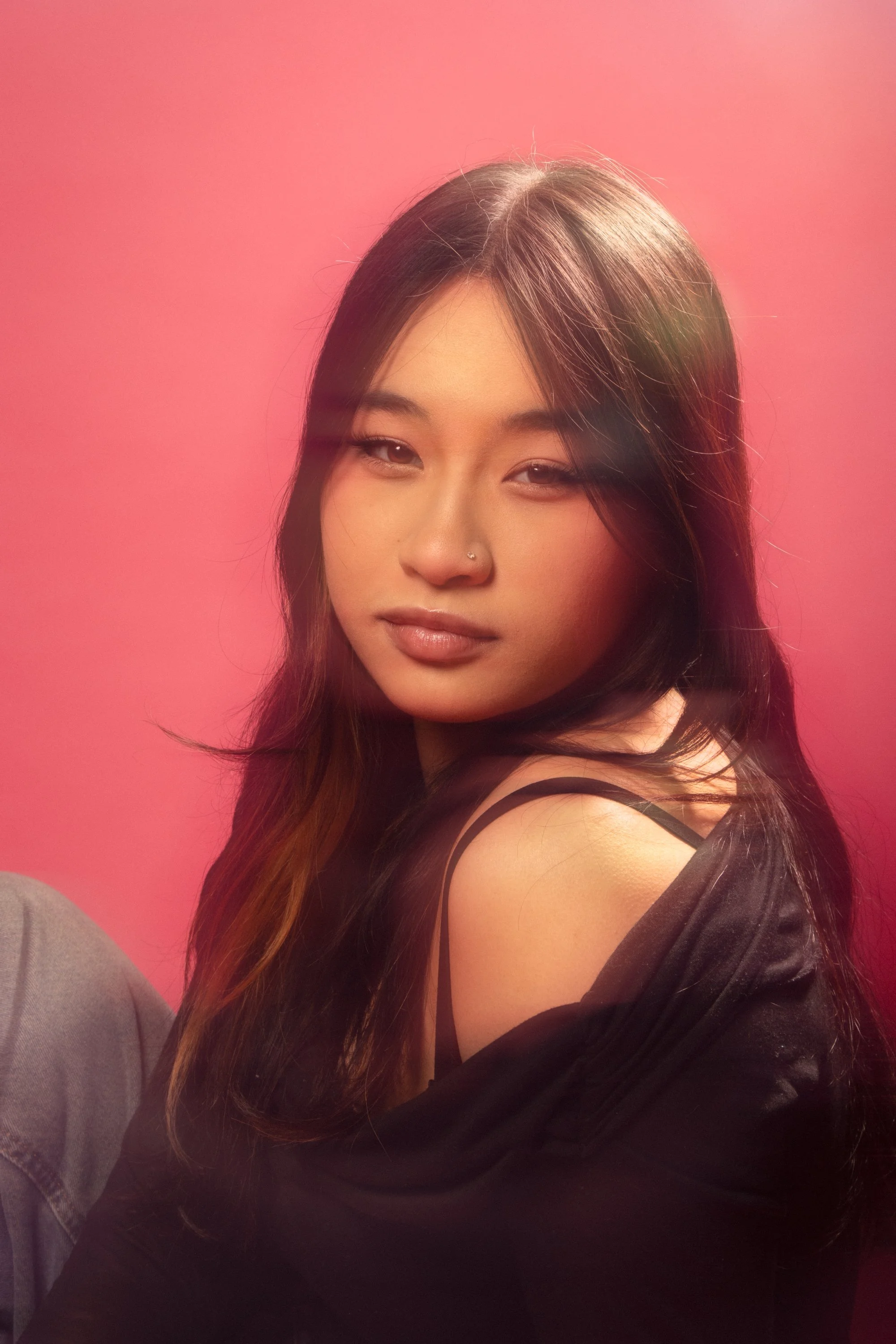 Portrait of a young woman with dark hair and a nose piercing, sitting against a pink background, wearing a black off-shoulder top.