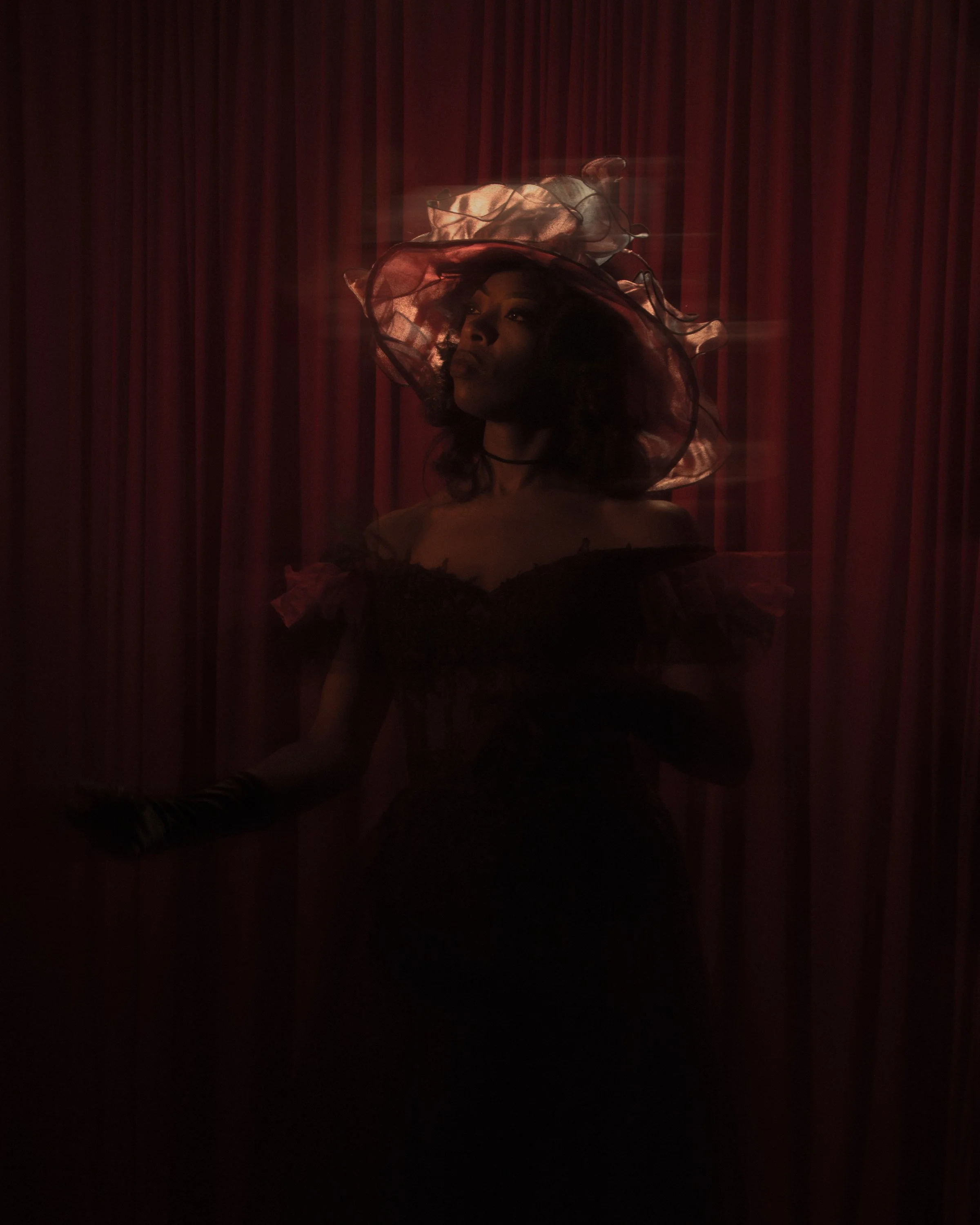 A woman sitting in front of red curtains wearing a large, translucent, ruffled dress and a matching wide-brimmed hat.