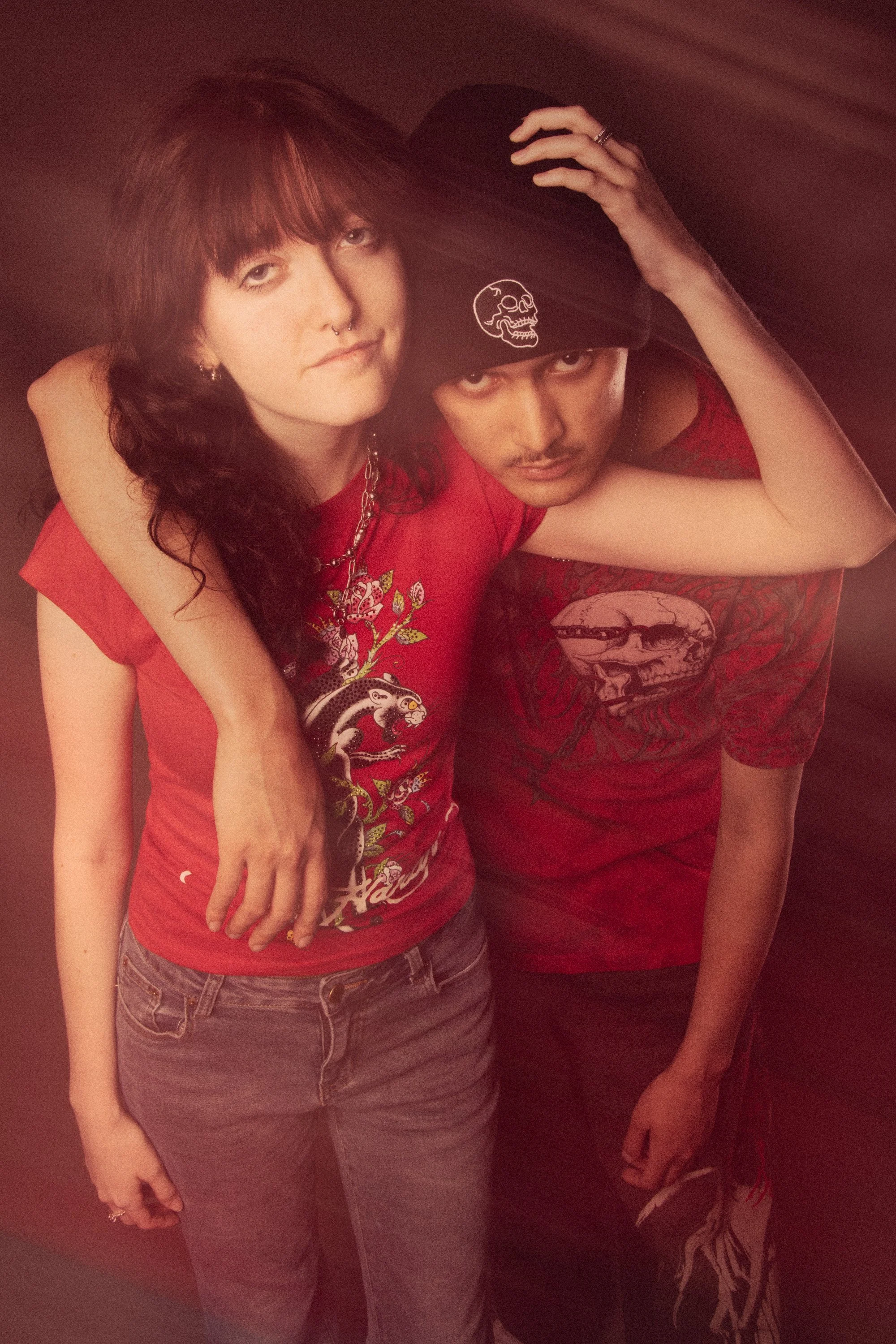 Two people close together, one wearing a red shirt with a dragon print and jeans, the other with a black beanie hat with a skull print, both with dark hair and edgy style, against a dark background.