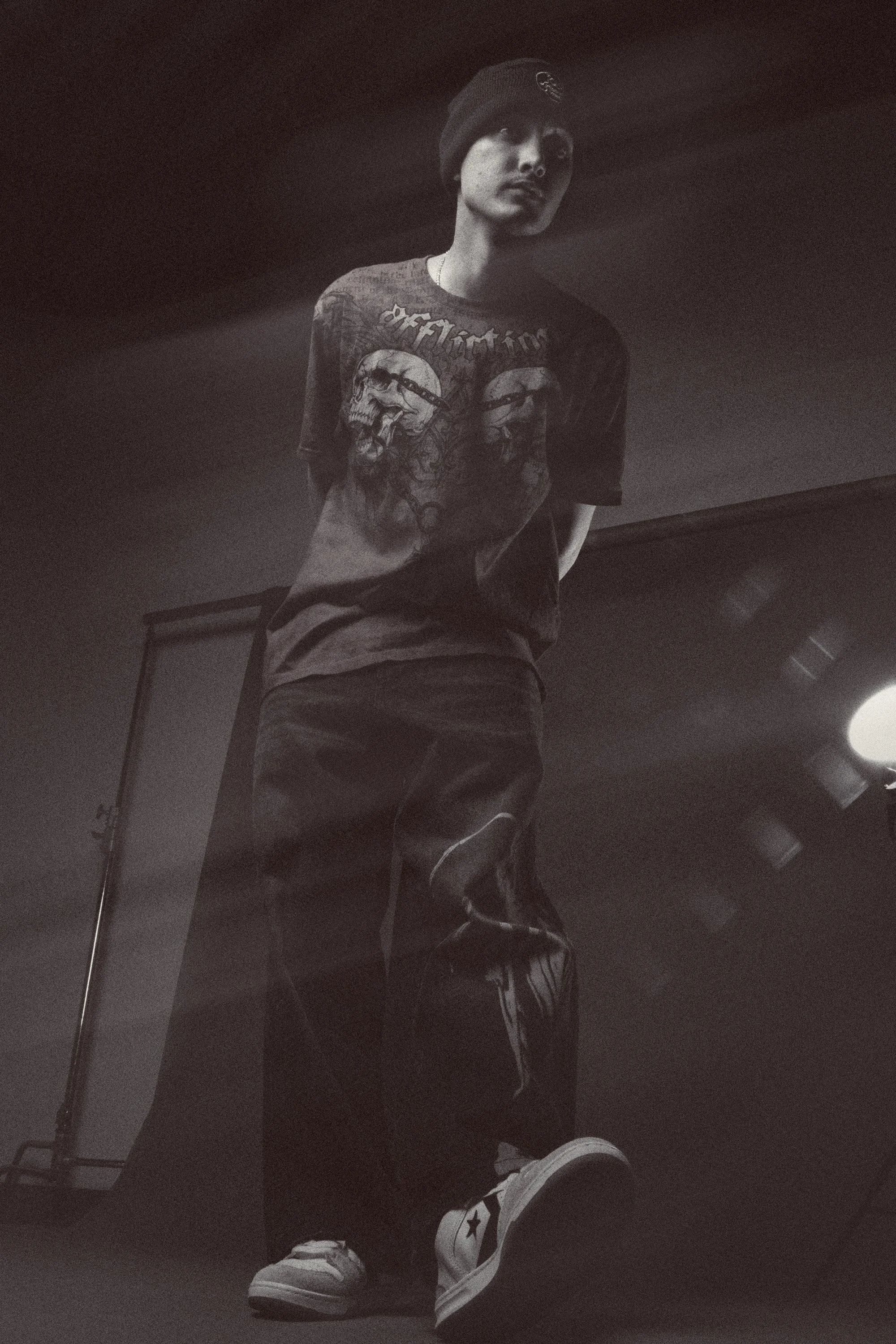 A person standing indoors wearing a black beanie, a graphic t-shirt with skull and text design, baggy pants, and high-top sneakers. The photo is black and white and taken from a low angle.