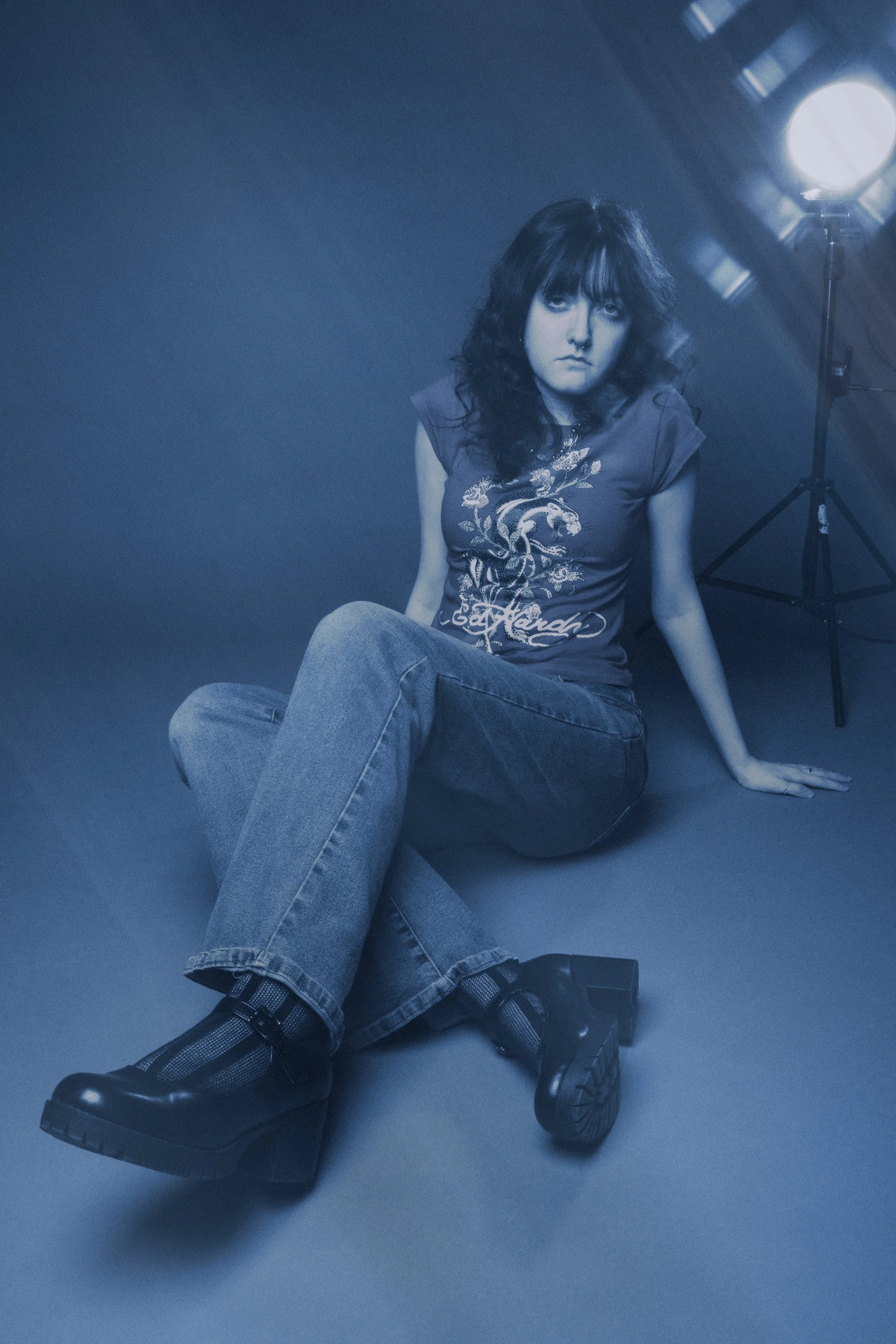 A young woman with dark, wavy hair and a septum piercing sits on the floor in a dimly lit room. She wears a black T-shirt with a dragon design and the word 'Loud,' along with loose jeans and chunky black platform shoes. A bright studio light on a tri