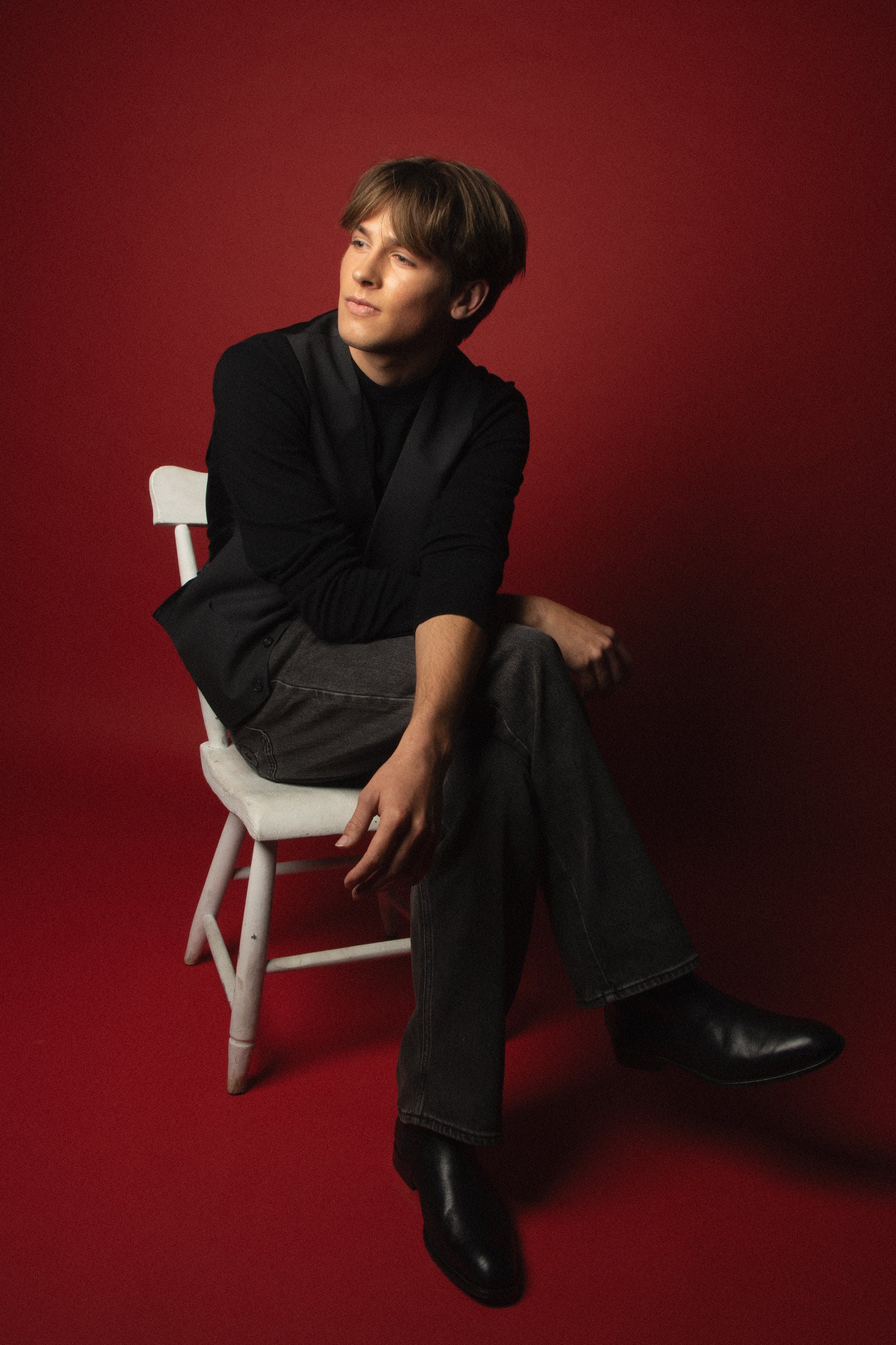 A person with brown hair and fair skin sitting on a white chair against a red background, wearing a black jacket and dark pants, with one leg crossed over the other and his arm resting on his knee.