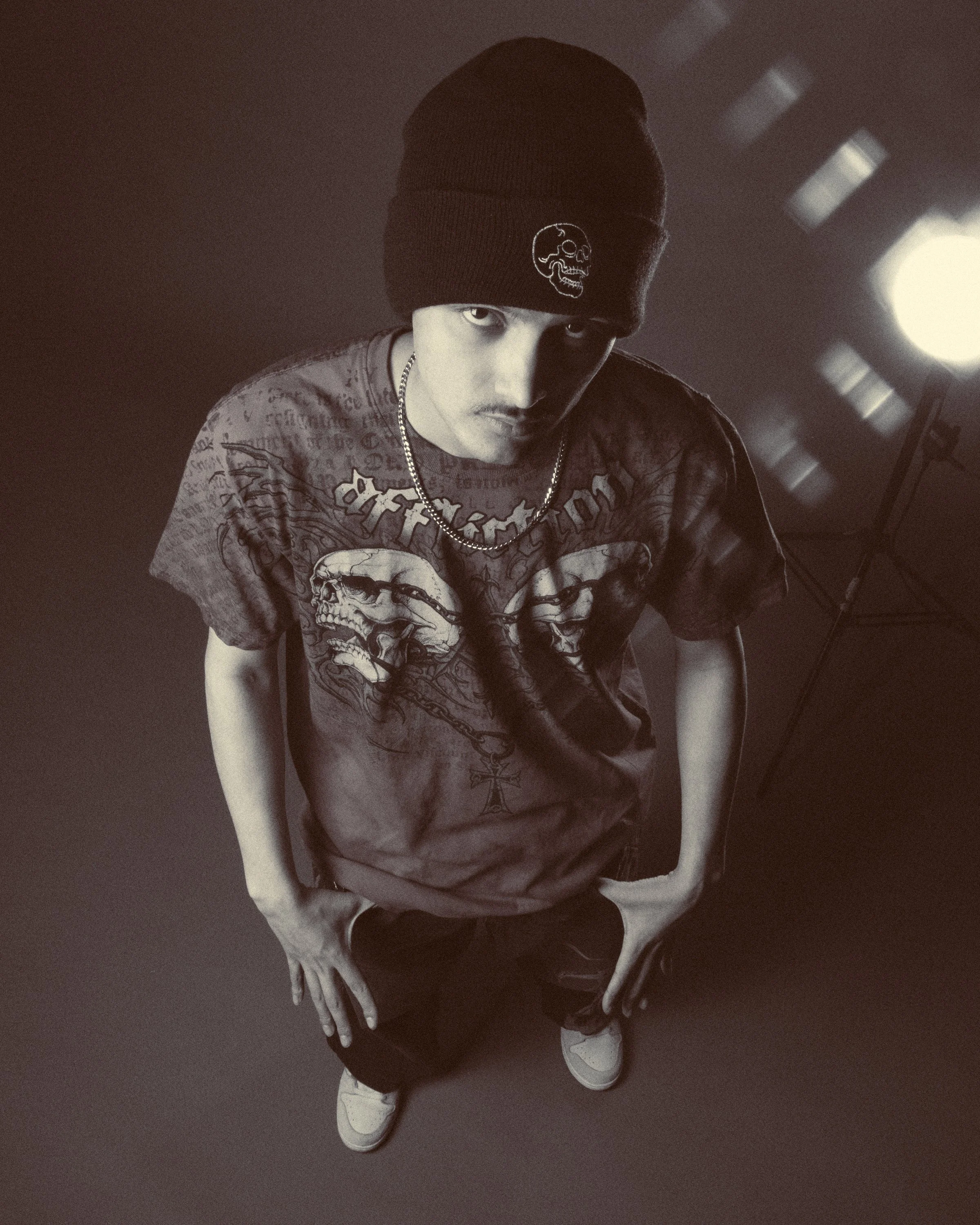 Young man wearing a black beanie with skull logo, graphic T-shirt with skulls, and white sneakers, standing on a dark floor, with his hands in his pockets, looking at the camera.