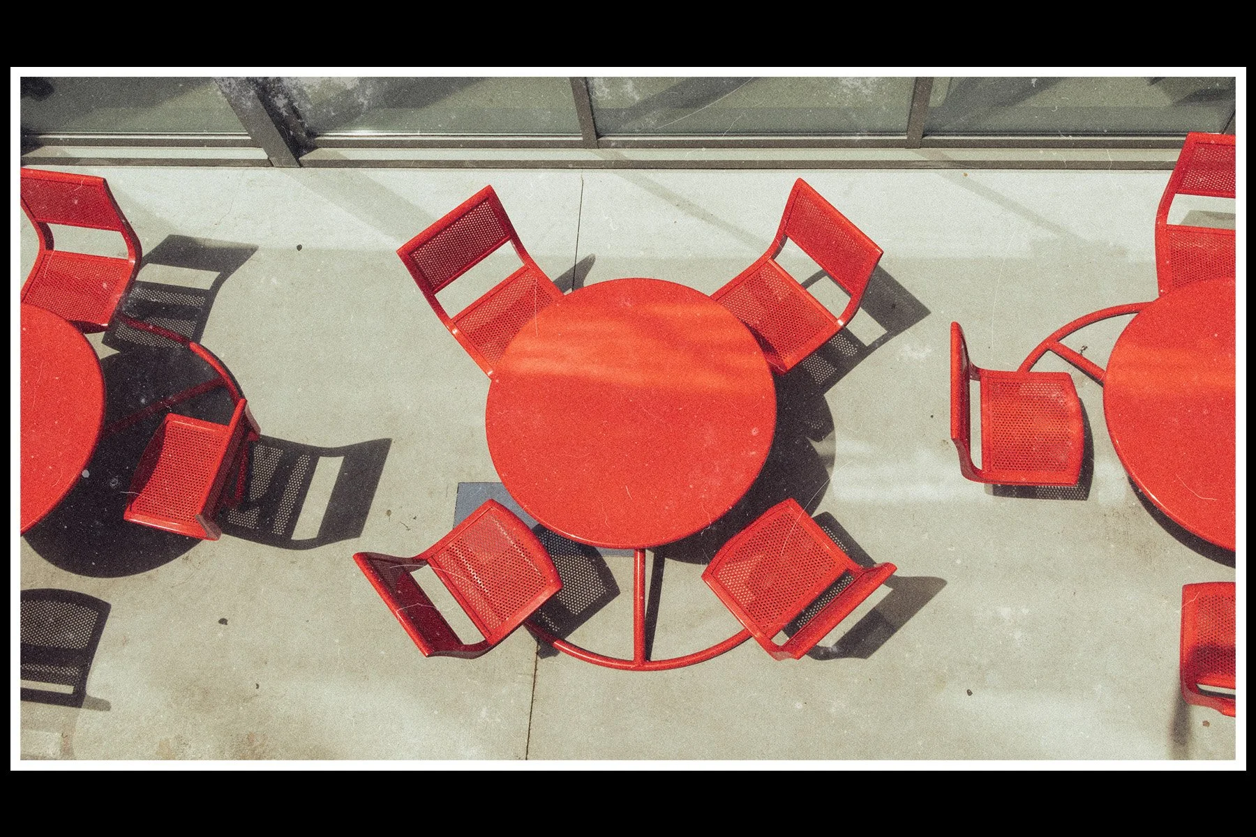 Red outdoor table with matching red chairs arranged around it, casting shadows on the concrete floor near a glass wall.