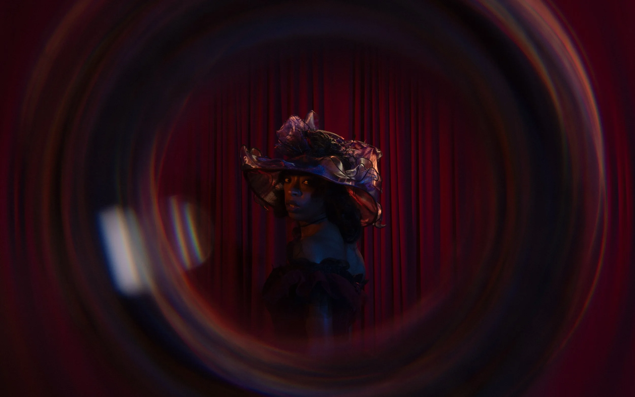 A woman wearing a large, translucent, ruffled hat with netting stands in front of red curtains, framed by a swirling, rainbow-colored vortex.