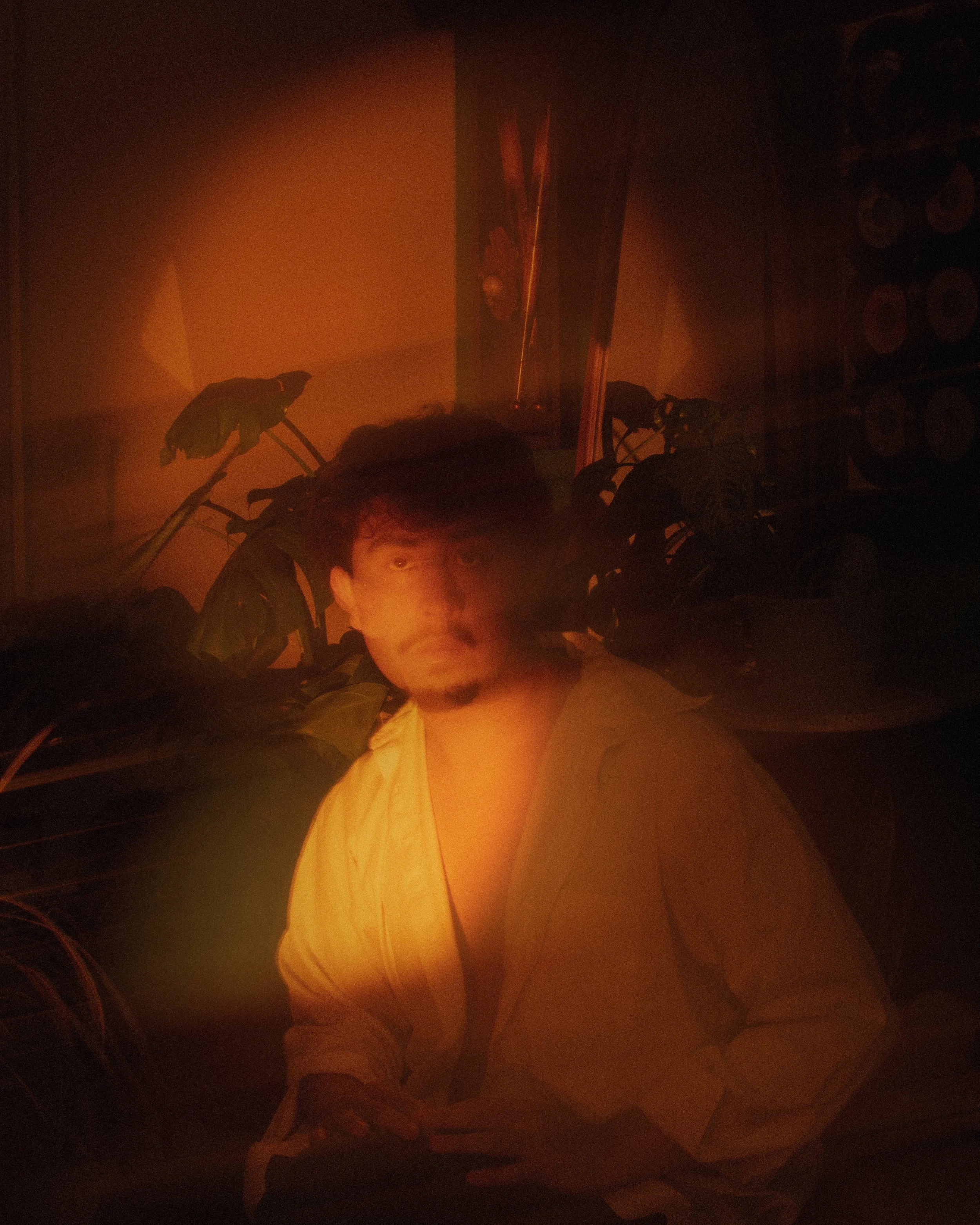 A young man with dark hair and a light beard, wearing a light-colored shirt, sits in a dimly lit room with orange lighting. He is surrounded by green plants and appears to be holding an object in his hands, with a blurry or reflective effect on his f