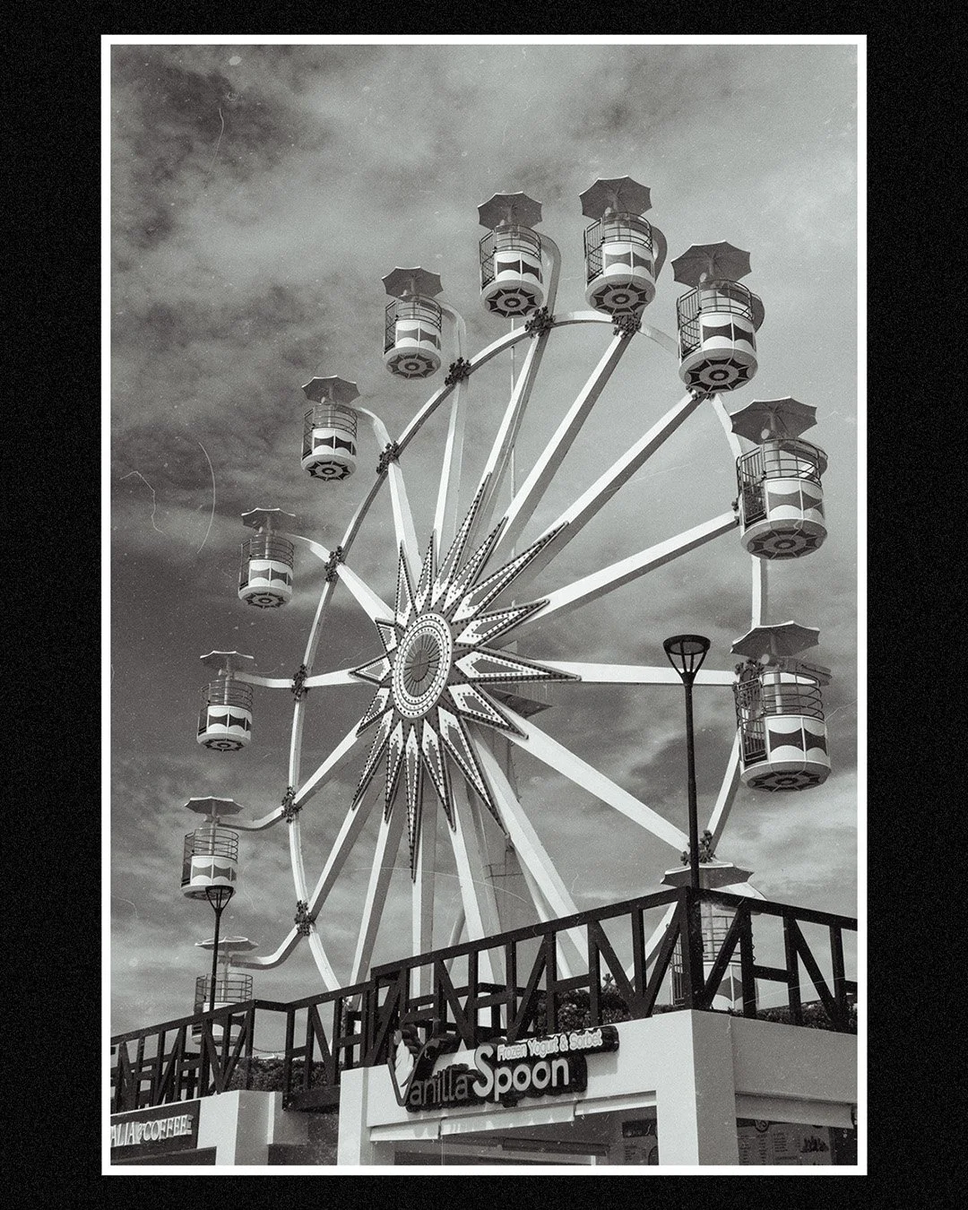 A black and white photo of a Ferris wheel at an amusement park, with a sign that reads "Vanilla Spoon" and a small part of another sign that reads "COFFEE."