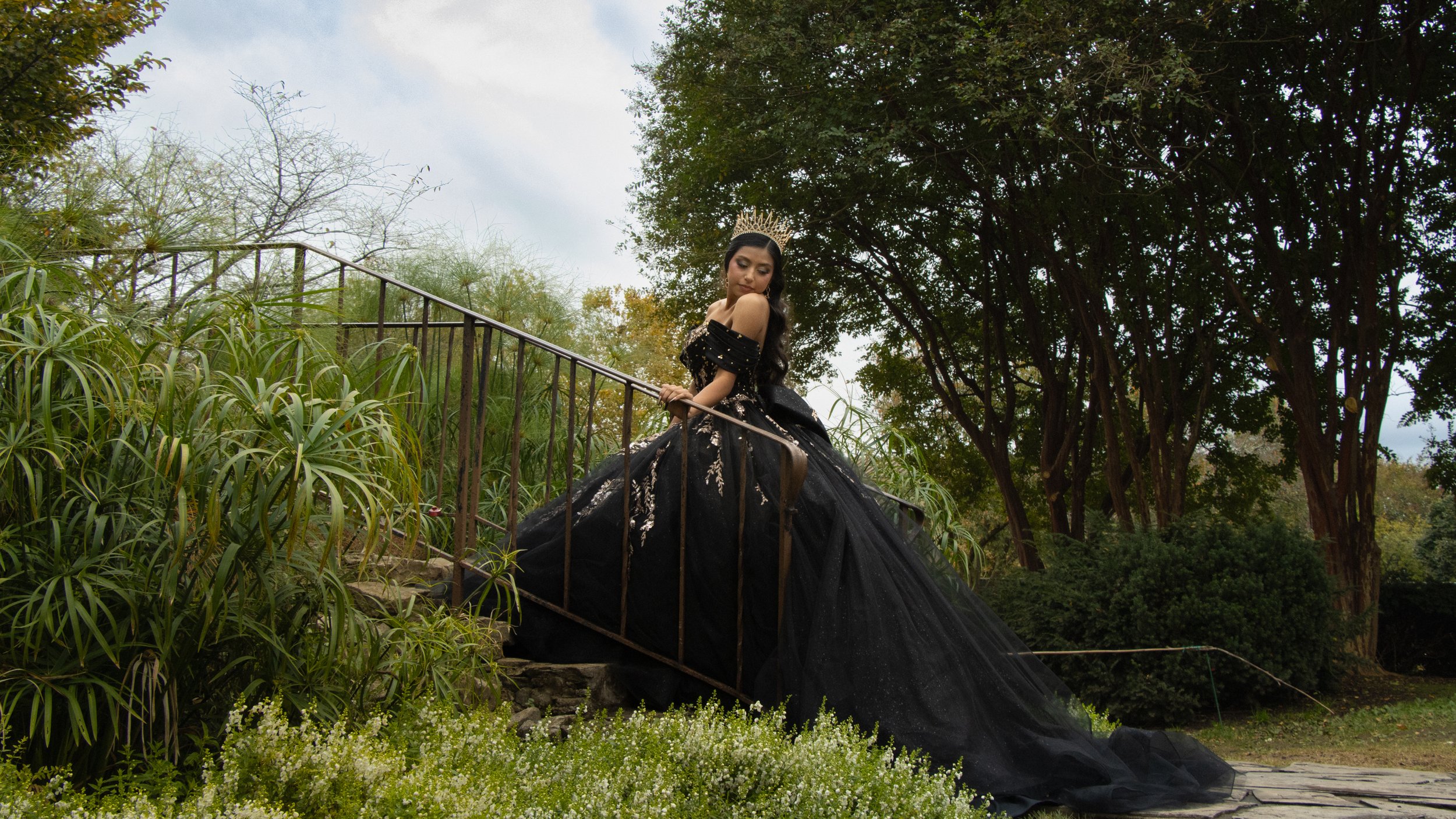 A woman in an elegant black gown with silver embroidery and a crown on her head, standing outdoors on a stairway amidst greenery and trees.