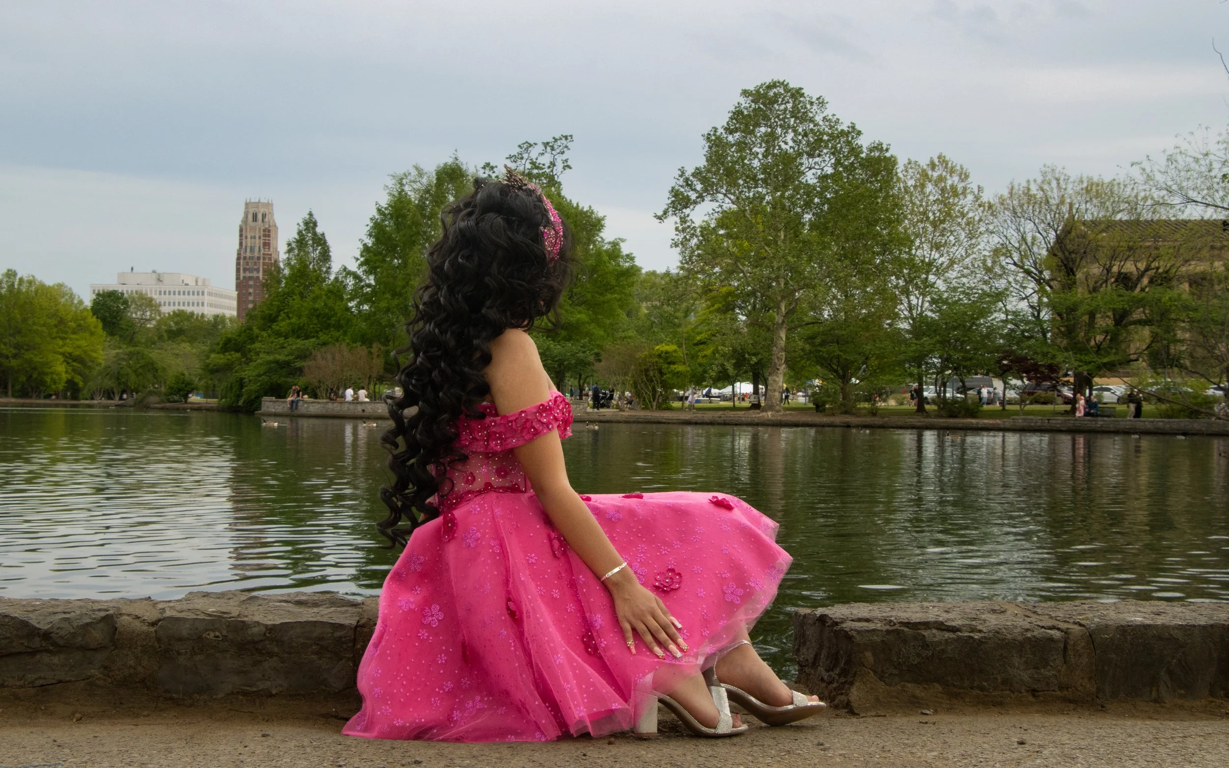 A girl wearing a pink dress with floral details, sitting on a rock beside a body of water in a park with green trees and high-rise buildings in the background.