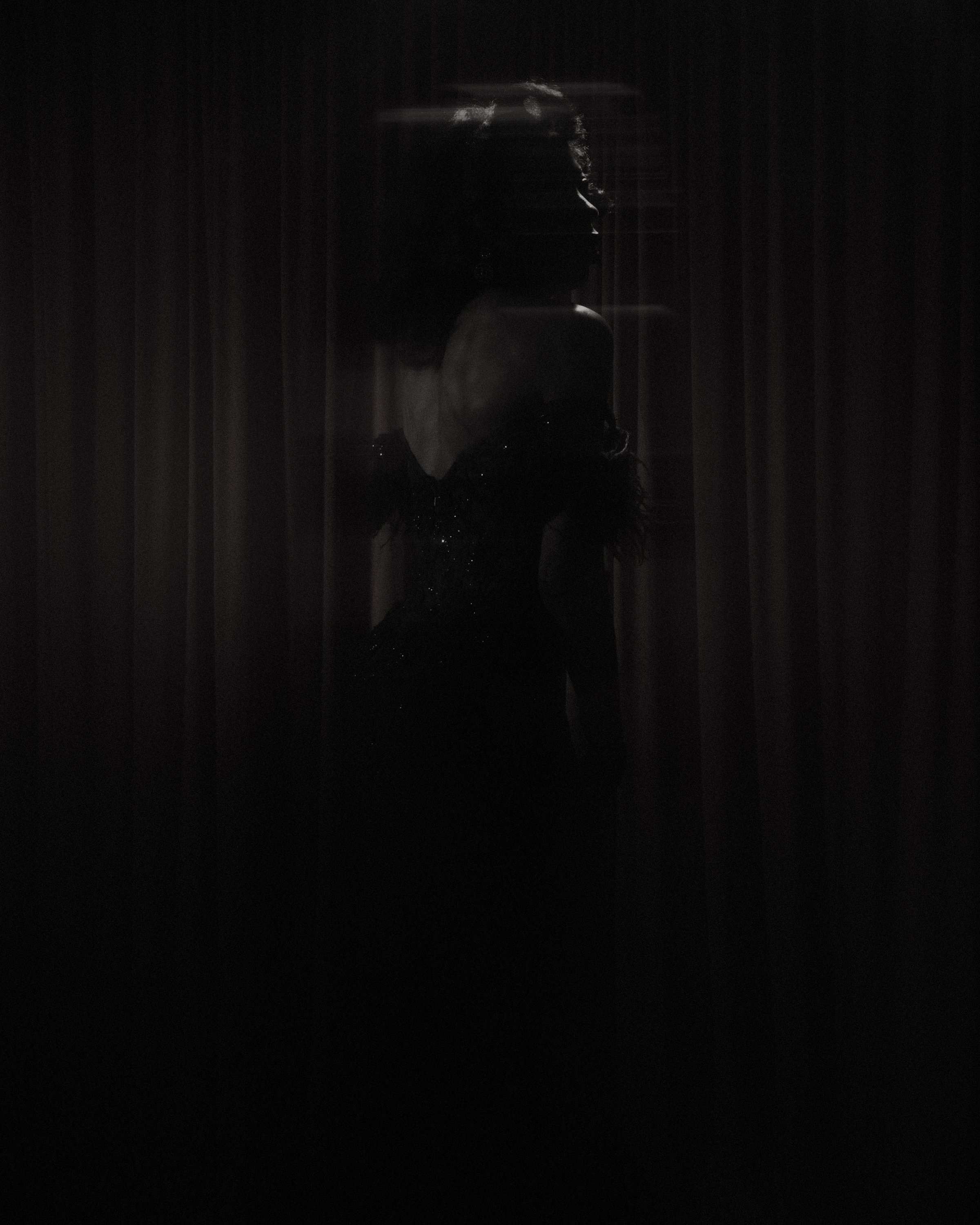 A dark silhouette of a woman standing in front of a curtain, with light behind her creating a shadowy effect.
