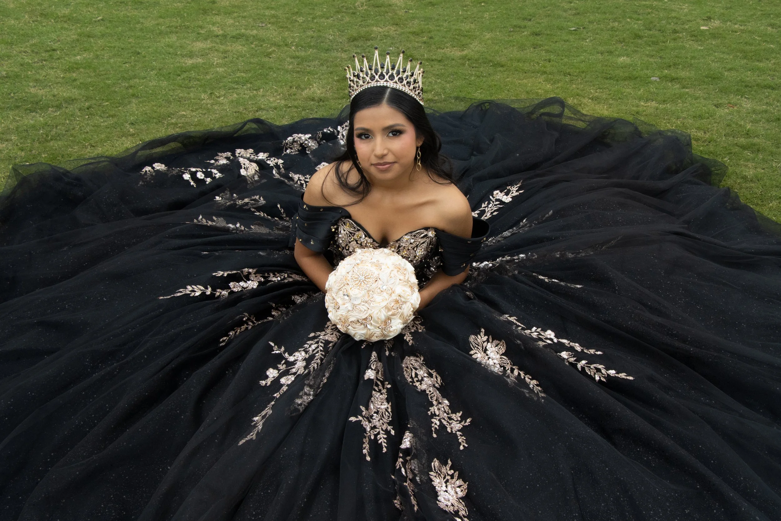 A woman wearing a black gown with silver embroidery, a silver crown, and gold earrings, sitting on the grass and holding a round bouquet of white roses.
