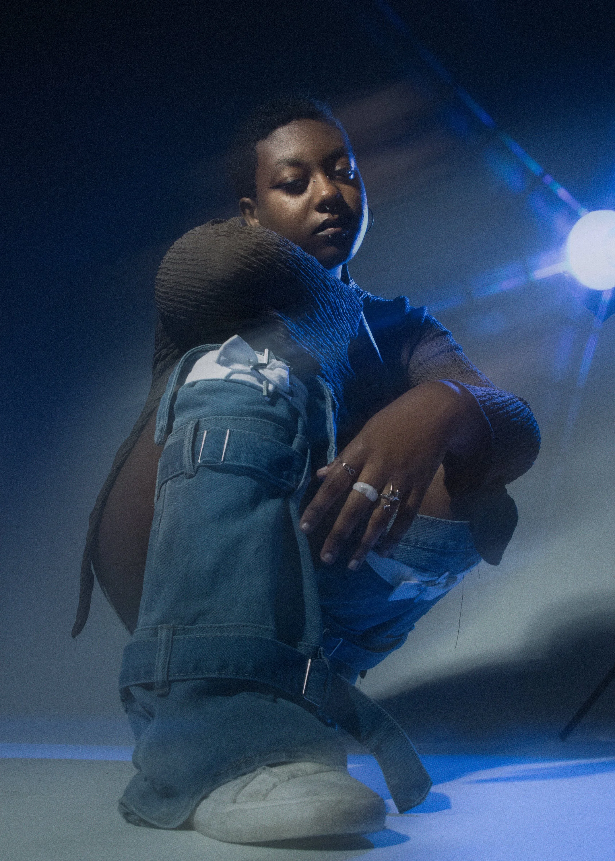 A person with dark skin and short hair squatting on the ground under blue lighting, wearing a textured brown sweater, blue jeans with a white cloth tucked into the waistband, and beige sneakers, with their arms resting on their knees.