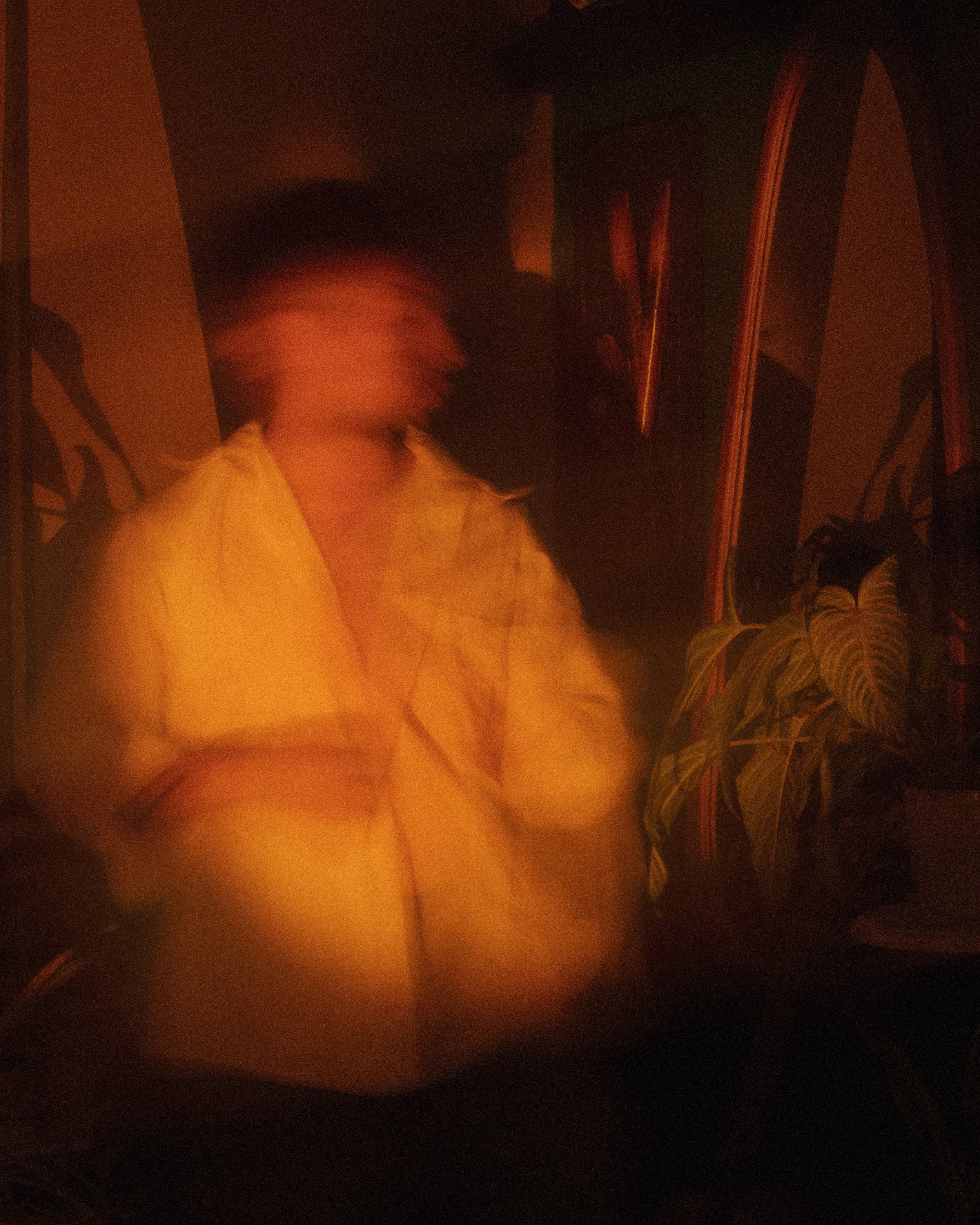 A person with short hair in a yellow jacket standing indoors next to houseplants, with a mirror and dark furniture in the background, illuminated by dim, warm lighting.