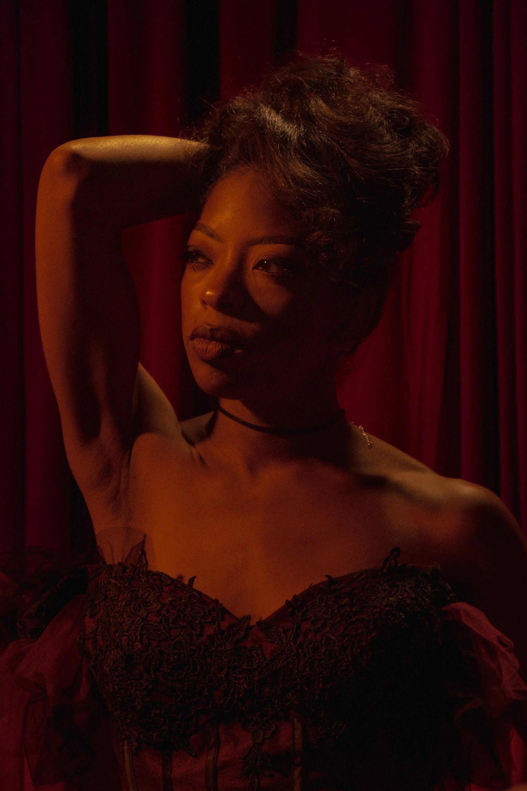 A woman with dark skin and curly hair poses in front of red curtains. She has her hand behind her head and is dressed in lace and tulle. The lighting is warm and dramatic.