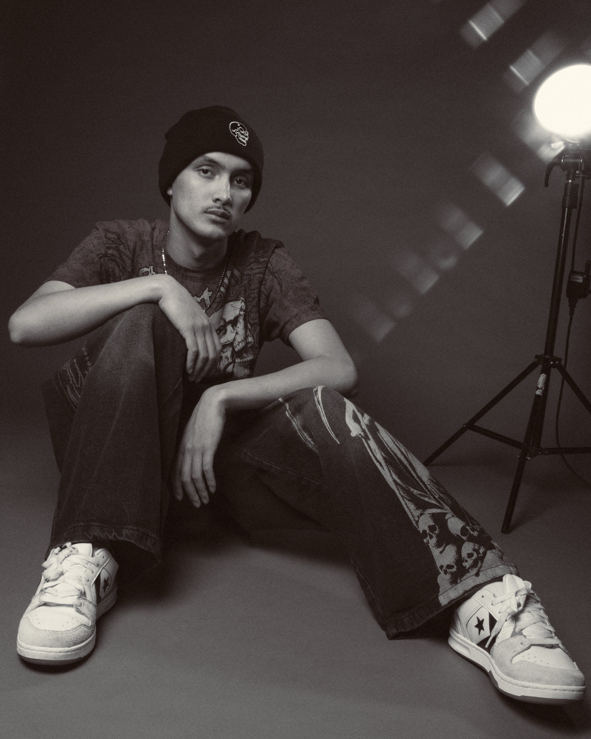 A young man sitting on the ground against a dark background, wearing a black beanie, graphic t-shirt, baggy jeans, and white sneakers, with studio lighting on the right side.
