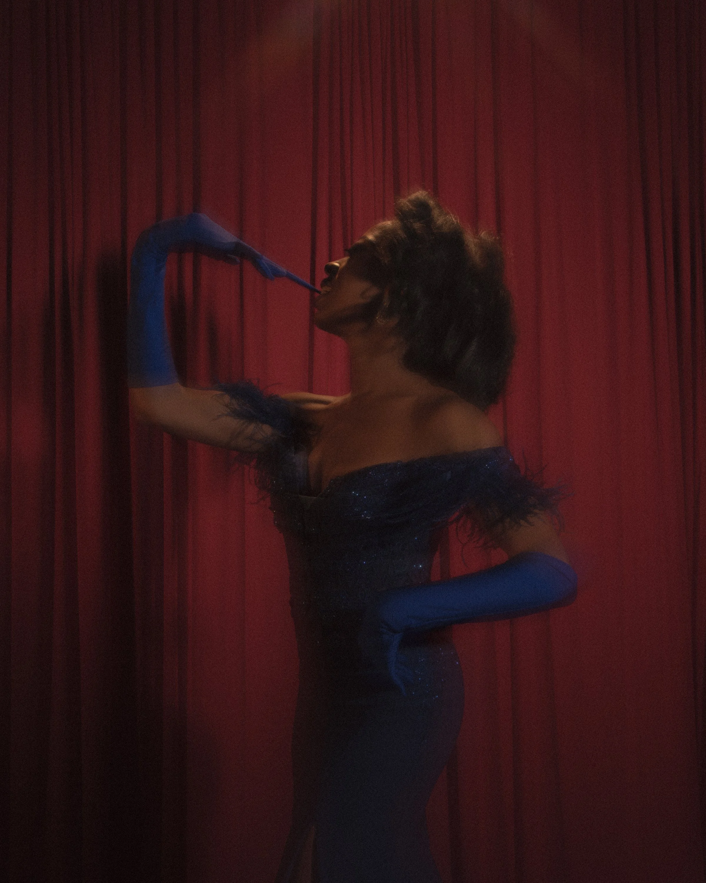 A woman in a sparkling blue dress and long blue gloves poses against a red curtain, holding a feathered fan near her face.