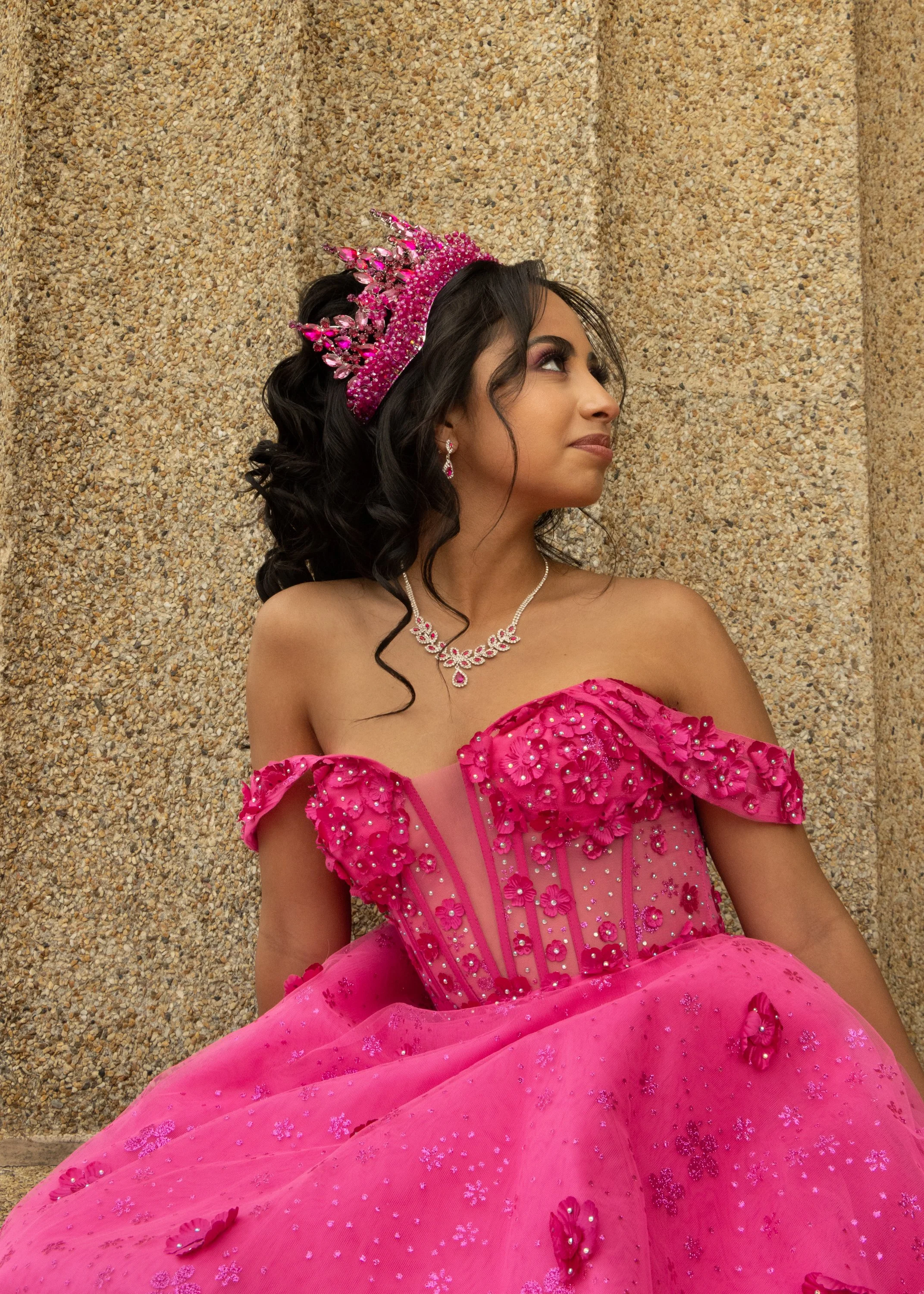A woman in a pink ball gown wearing a crown and jewelry, sitting against a textured wall, looking to the side.