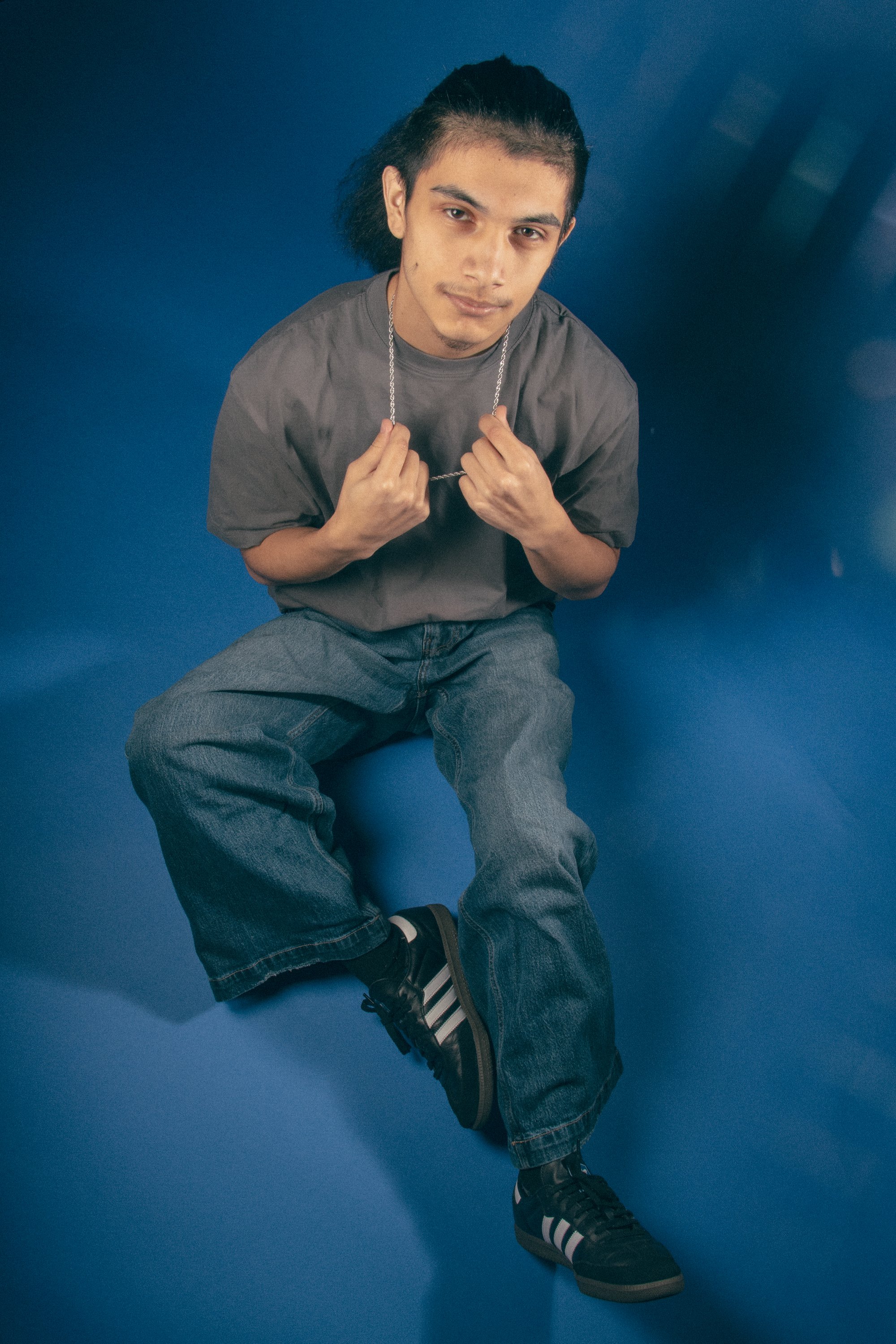 Young man with long hair, sitting against a blue background, wearing a grey t-shirt, baggy jeans, and black Adidas sneakers, holding a silver chain with both hands, looking at the camera.