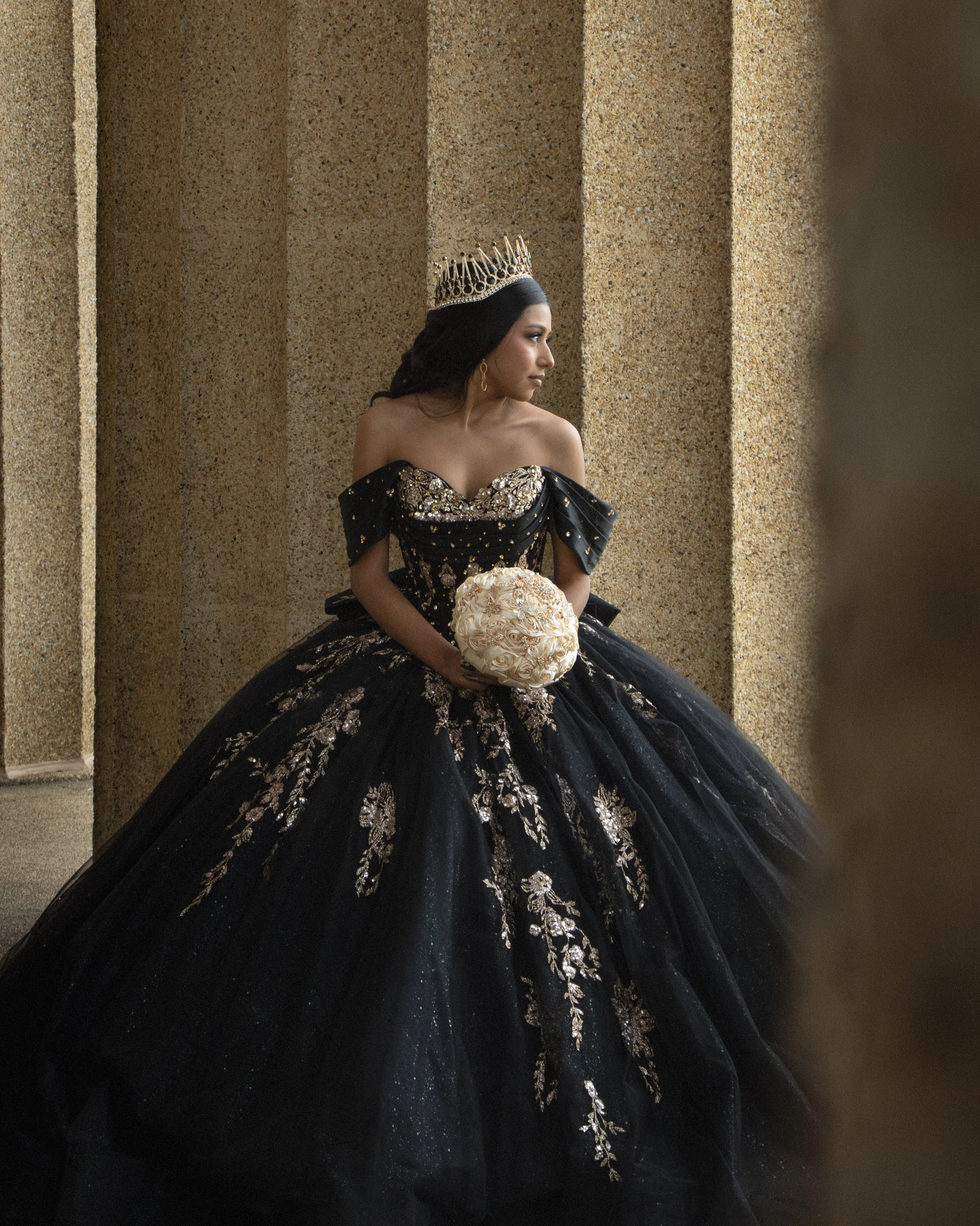 A woman wearing an elaborate black gown with gold embroidery and a tiara, holding a bouquet of white roses, seated against a textured beige wall.