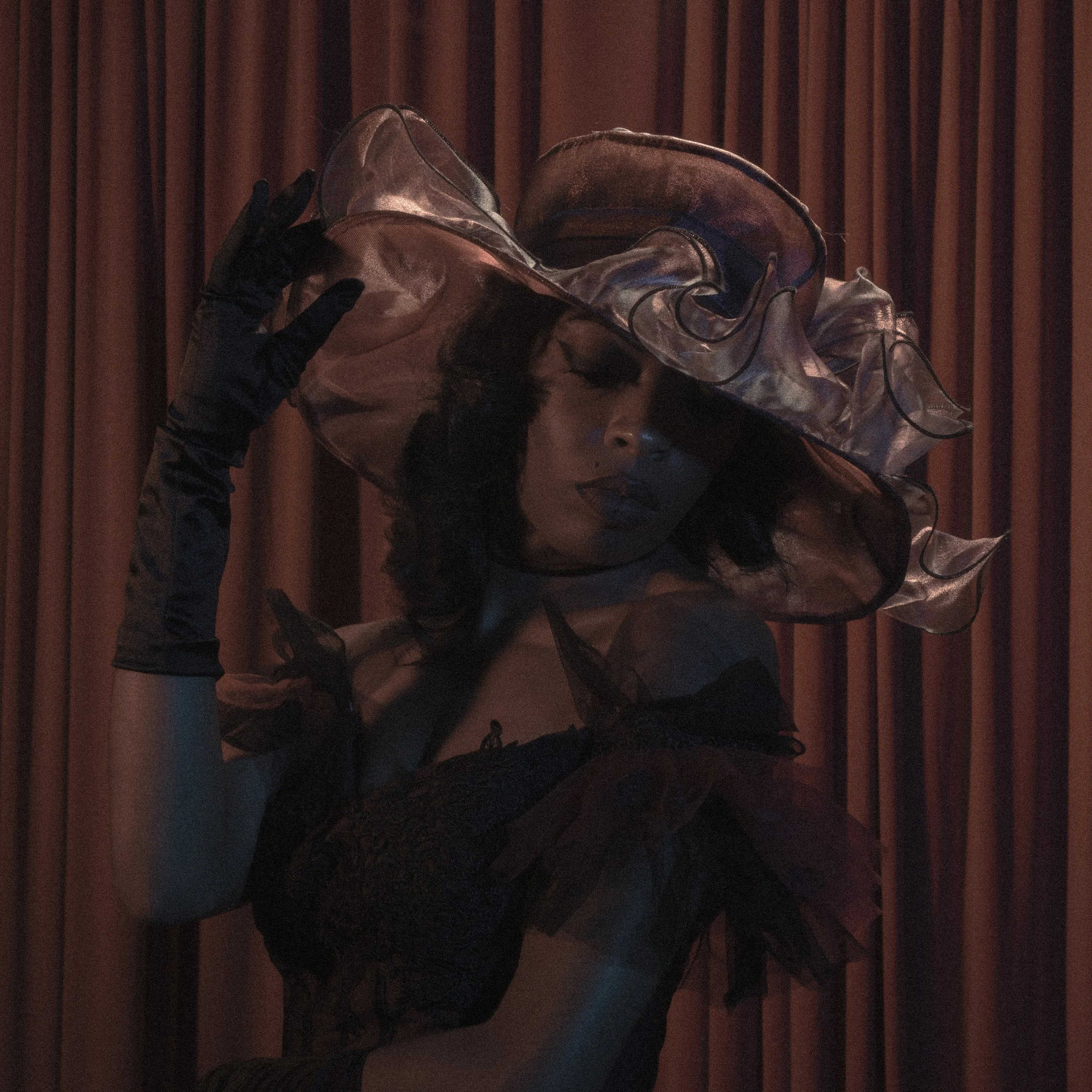 A person wearing black gloves is holding a clear plastic face shield, with brown curtains in the background.