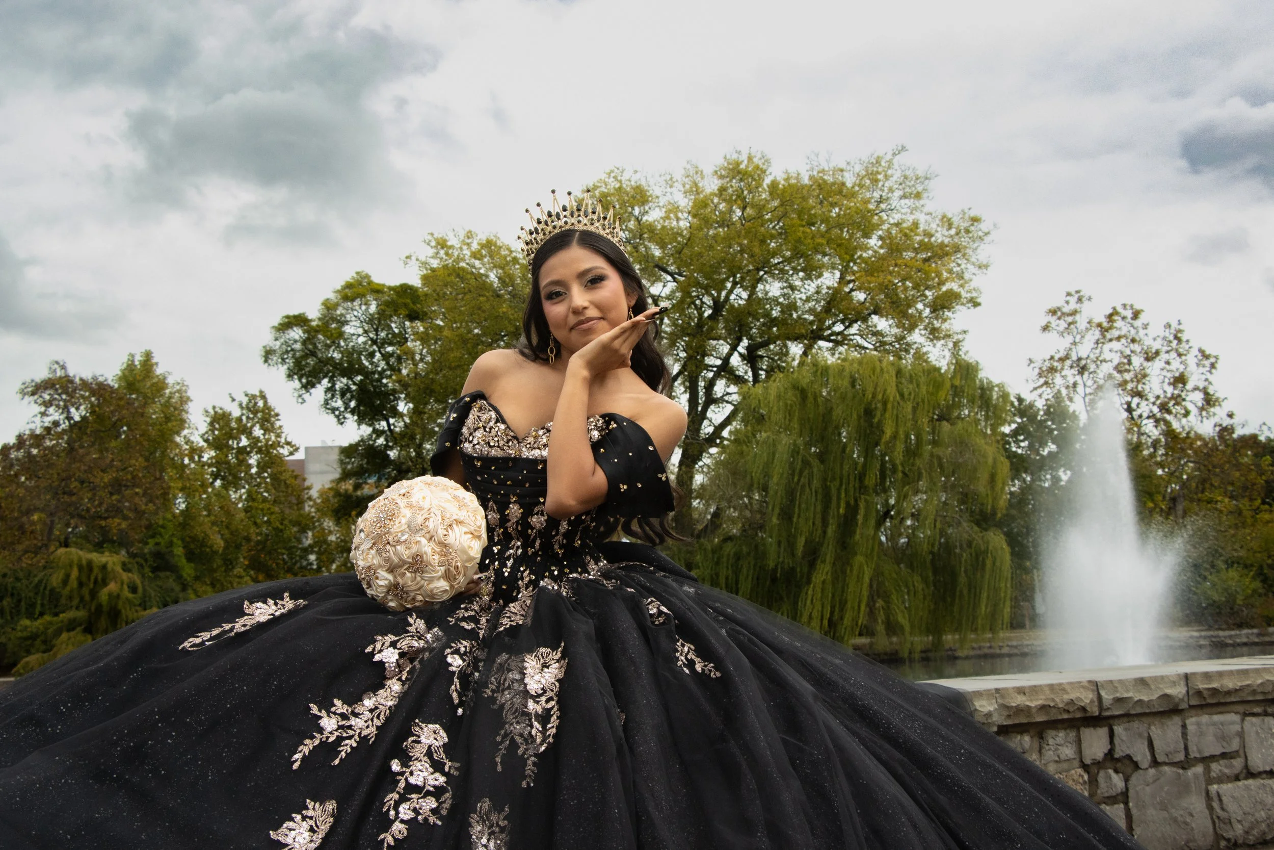 A woman in an elegant black and gold gown with intricate embroidery and a crown, sitting outdoors near water and trees during daytime.