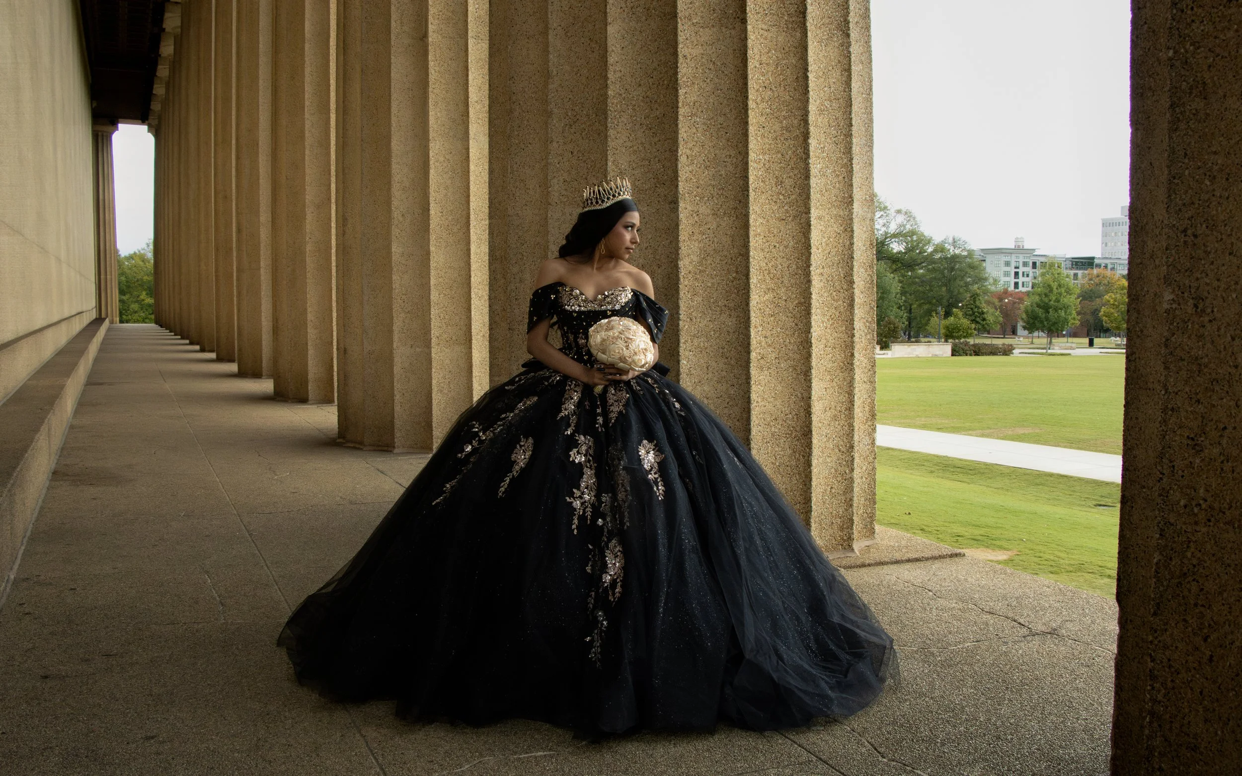 A woman wearing a black, off-the-shoulder ball gown with gold and silver embellishments, standing under a colonnade, holding a bouquet of flowers, and wearing a crown, outdoors near a park and city buildings.