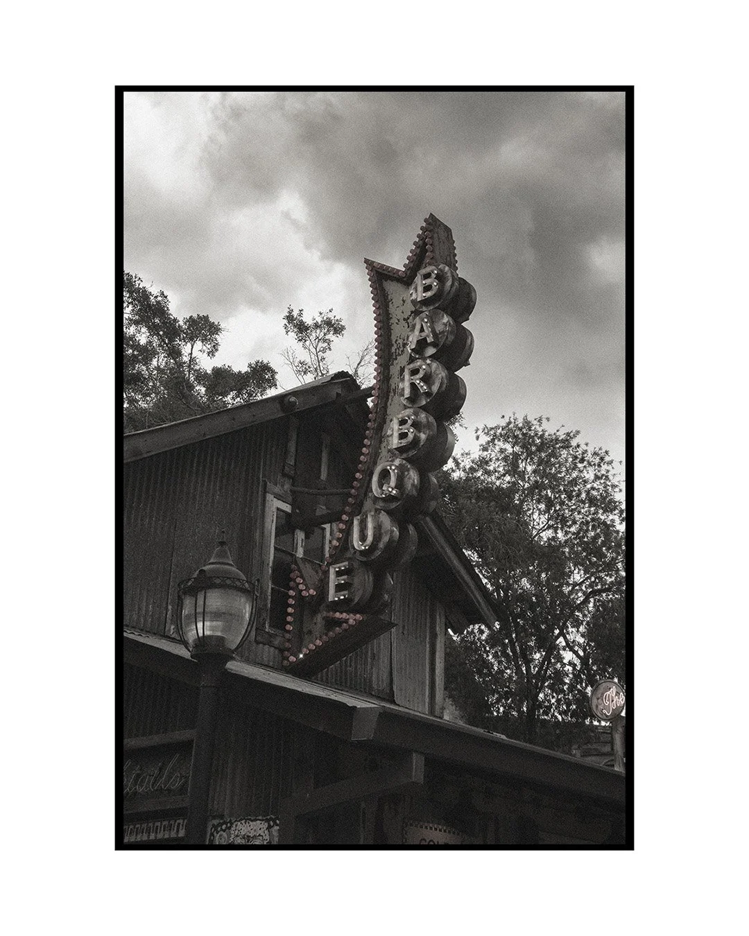 A vintage-style vertical sign with illuminated letters spelling 'BARBQUE' outside a rustic wooden building with a lantern, under a cloudy sky and trees.