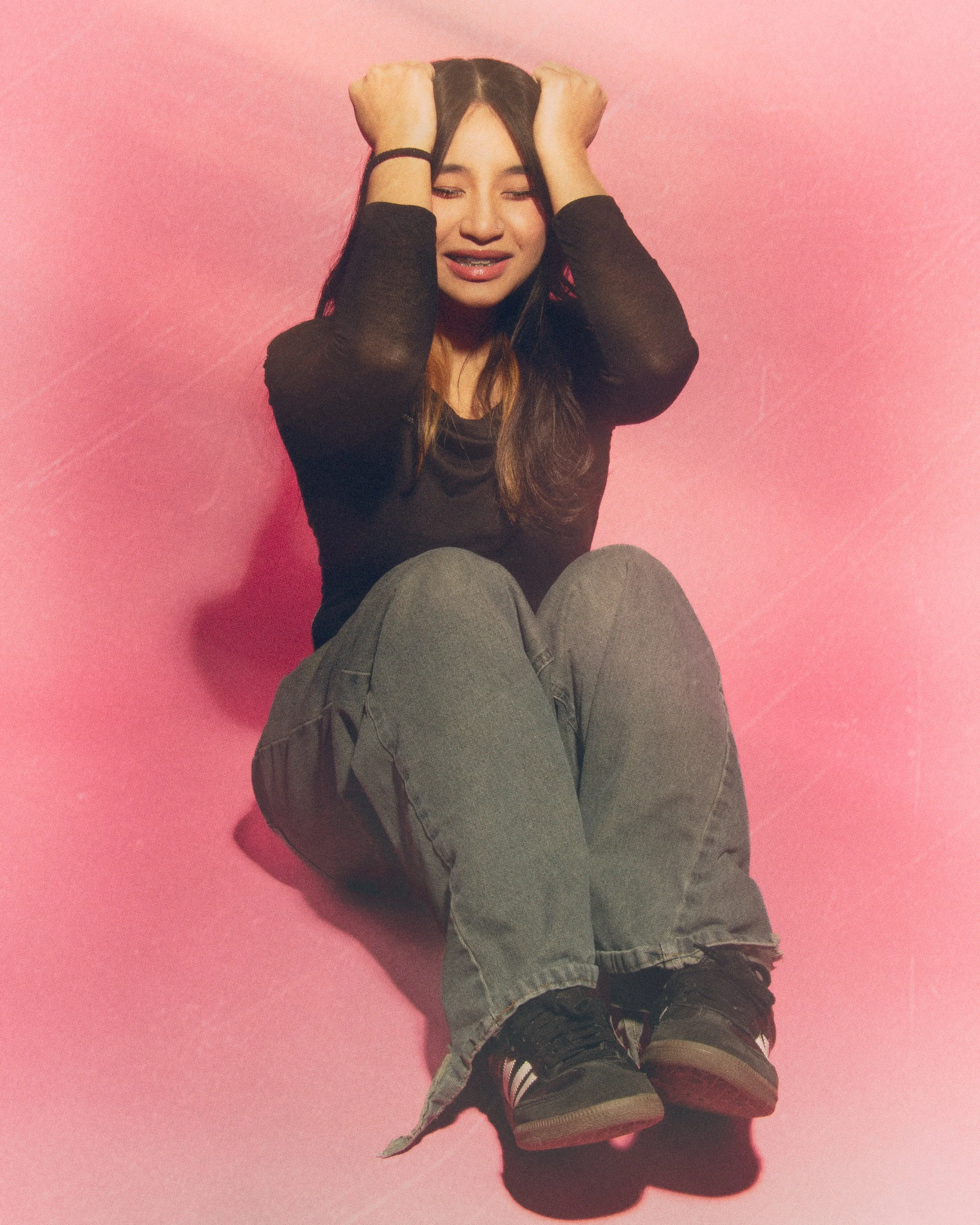 A young woman with long dark hair sitting against a pink wall, wearing black long-sleeve top, gray jeans, and black shoes, with her hands holding her head, looking distressed.